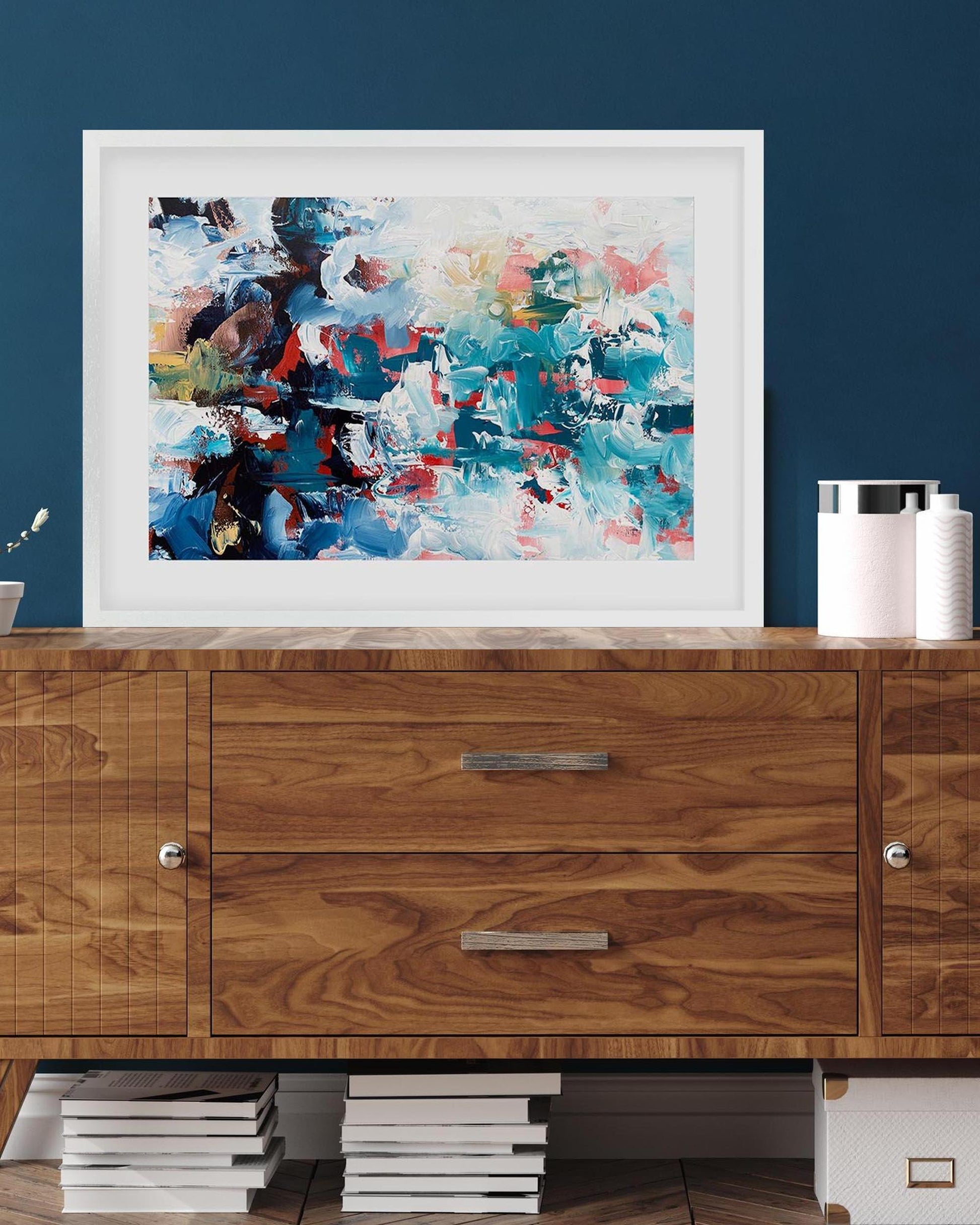 Abstract painting in a white frame on a wooden cabinet against a blue wall.