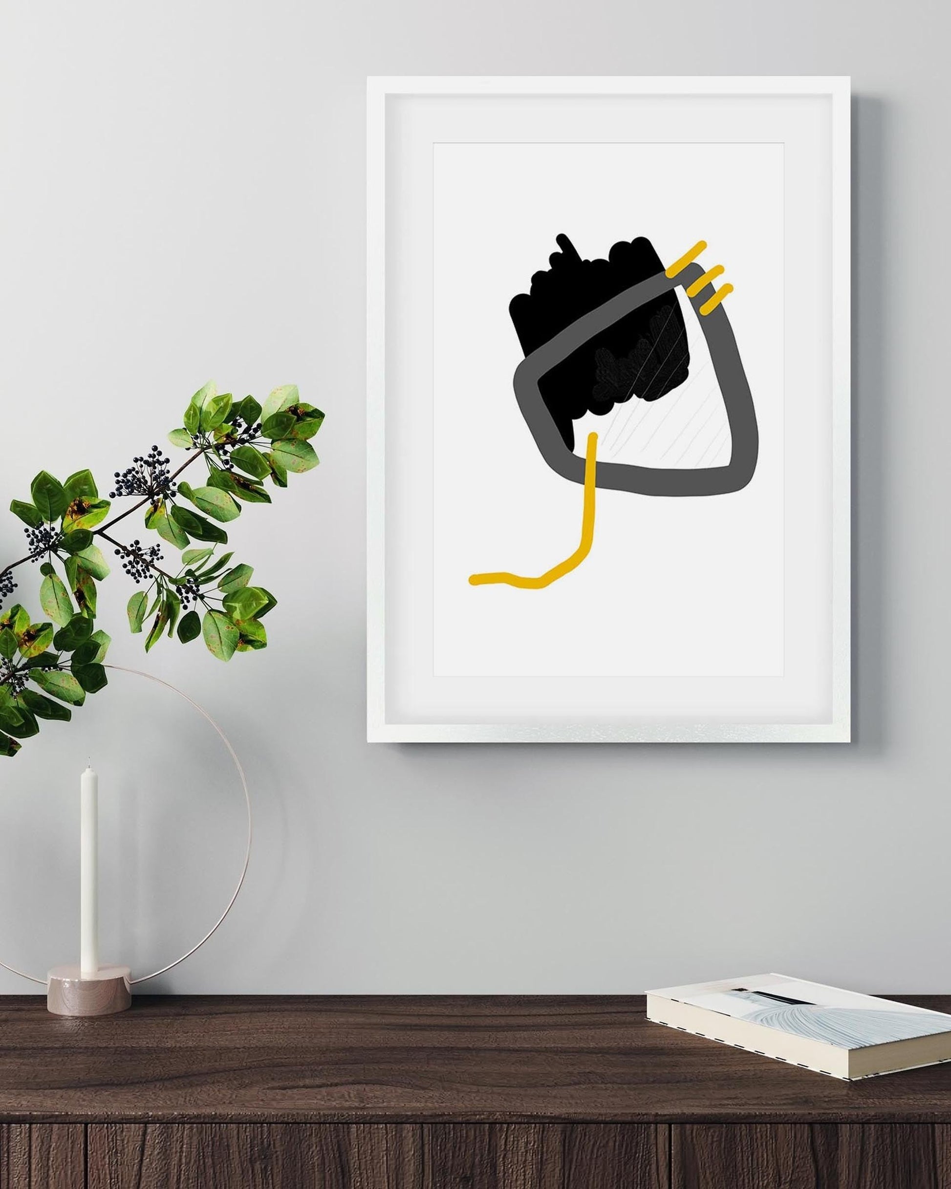 Framed abstract art piece on a wall with a plant and books below.