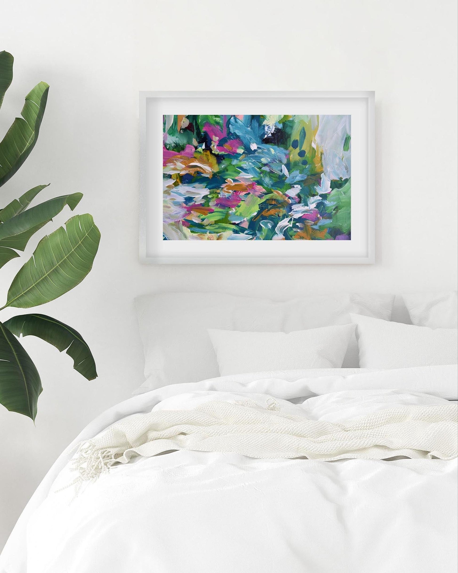 Framed abstract painting on a wall above a bed with a plant to the left.