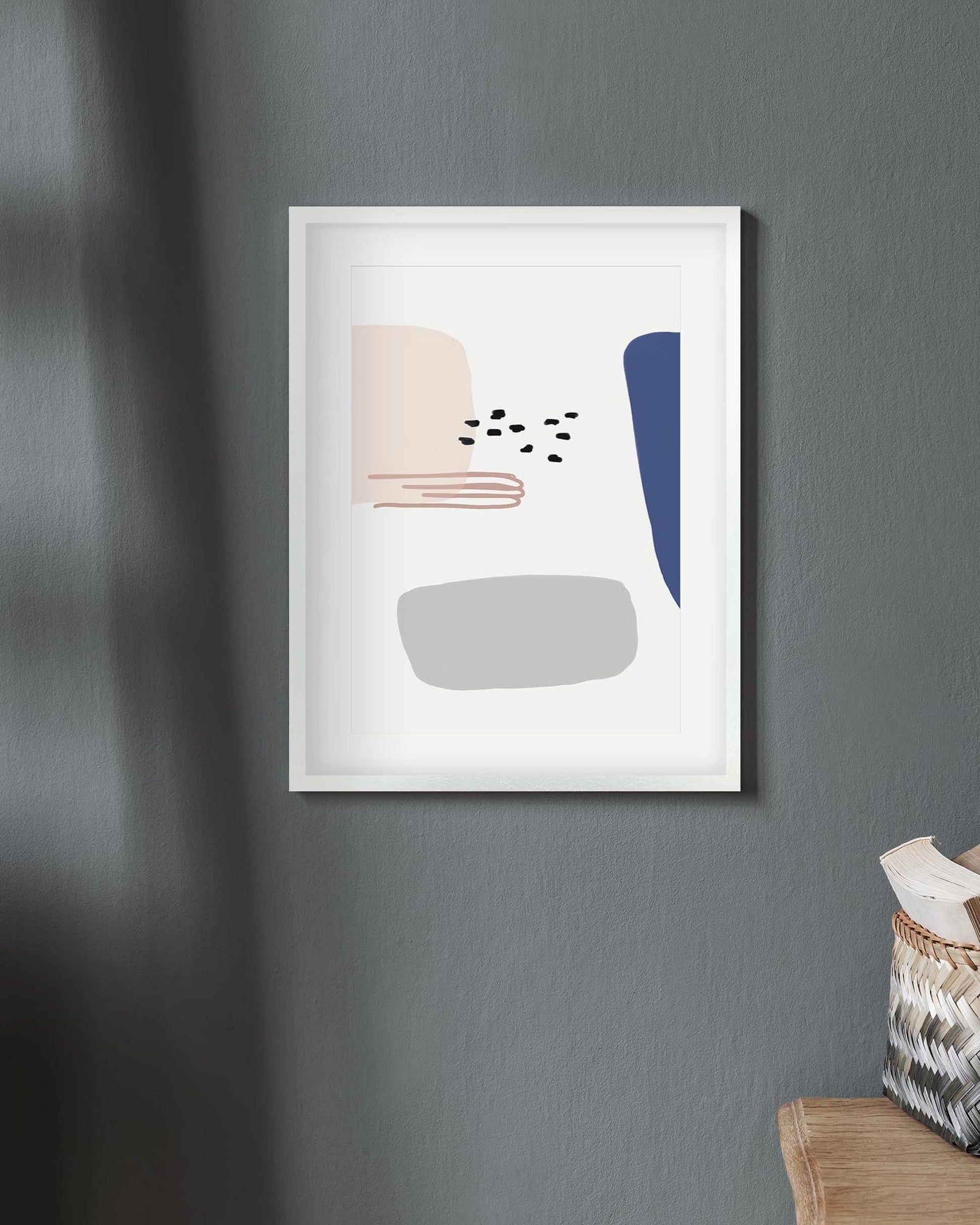 Framed abstract art print on a gray wall with a basket of books on a wooden surface.