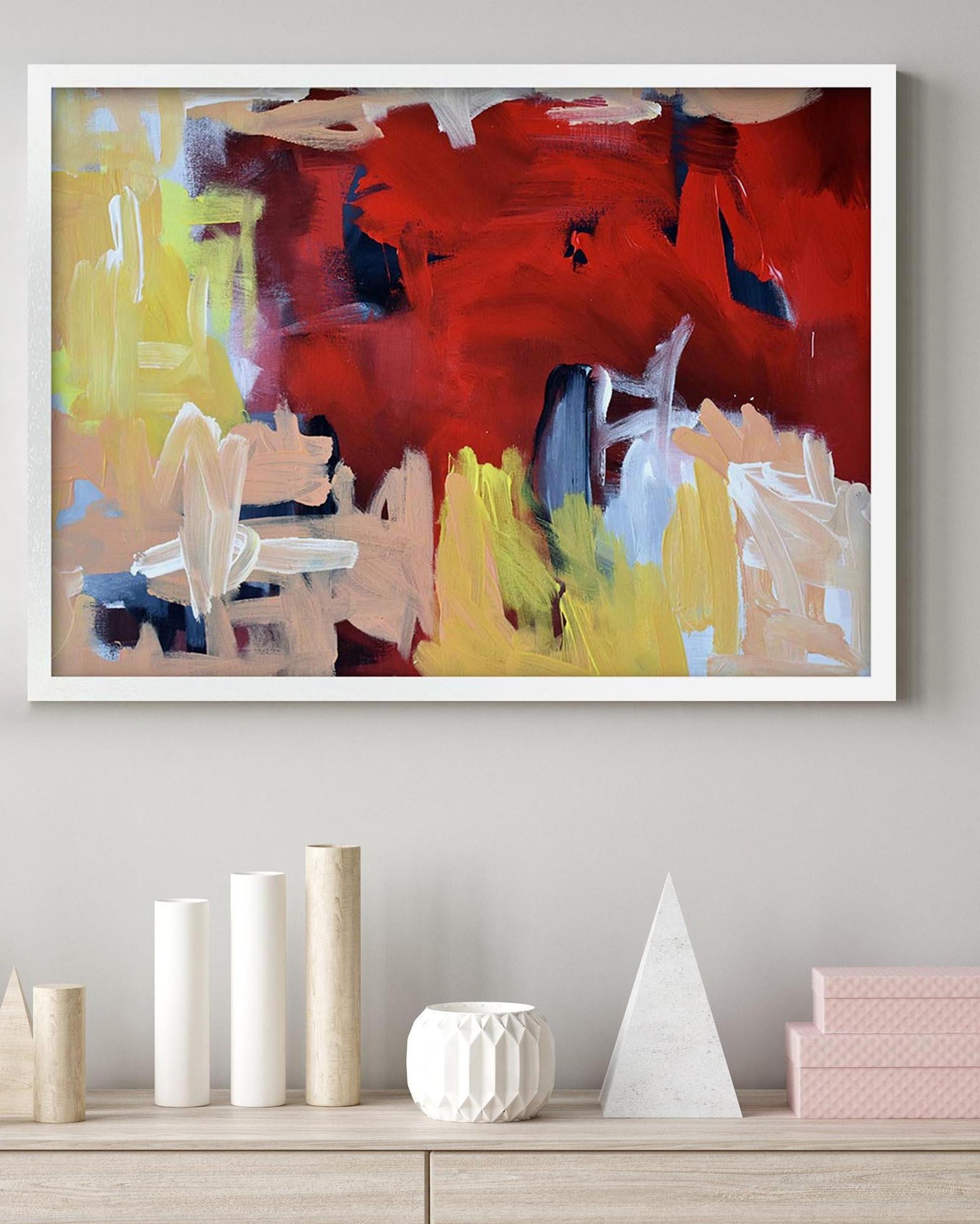Abstract painting in a white frame on a wall above a shelf with decorative items.