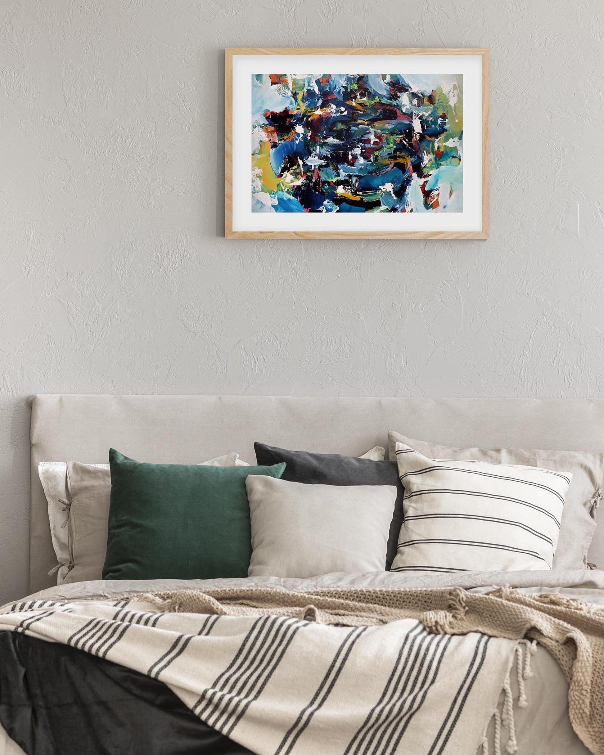Framed abstract painting above a bed with pillows and a blanket.