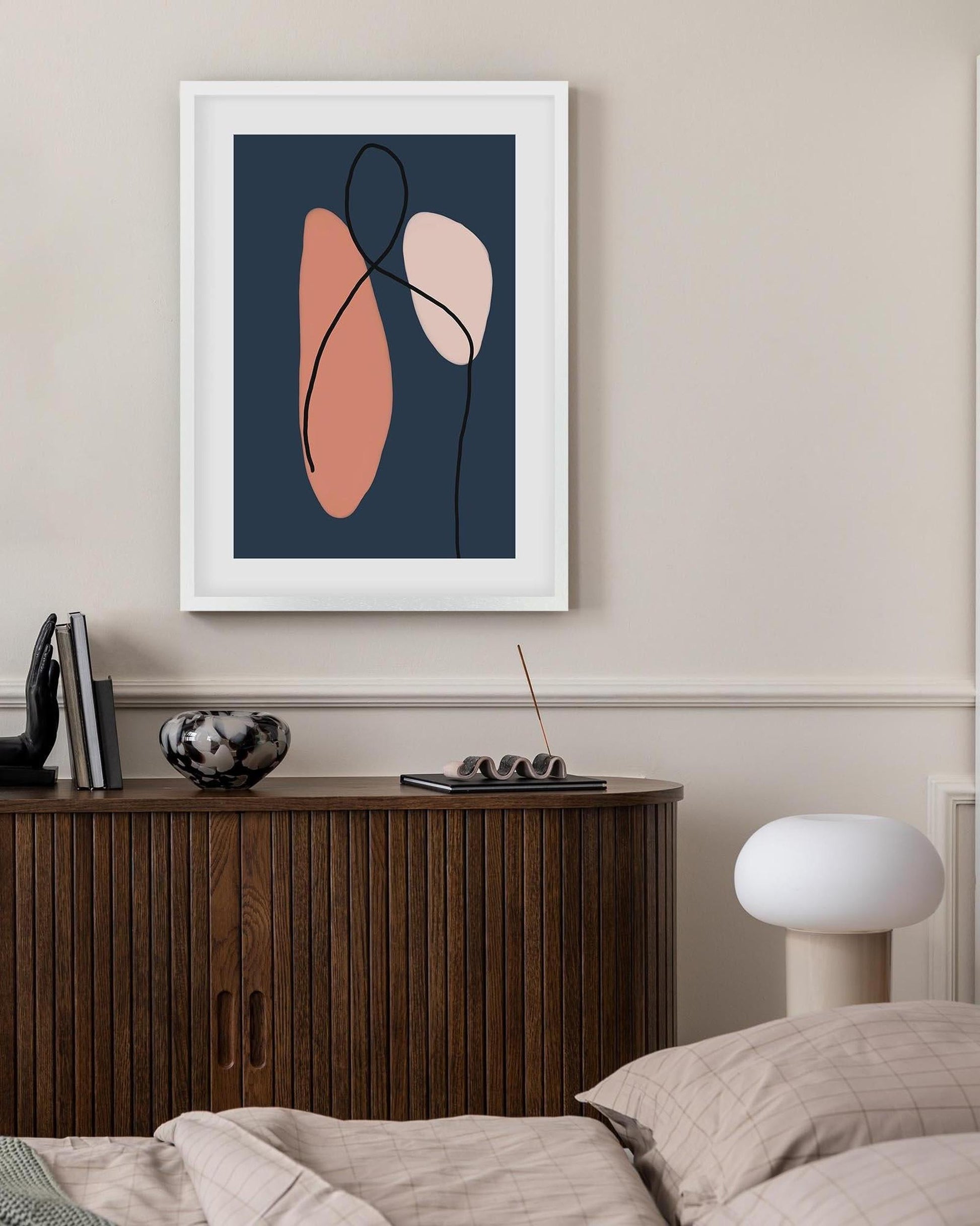 Framed abstract art piece on a wall above a wooden cabinet in a bedroom.