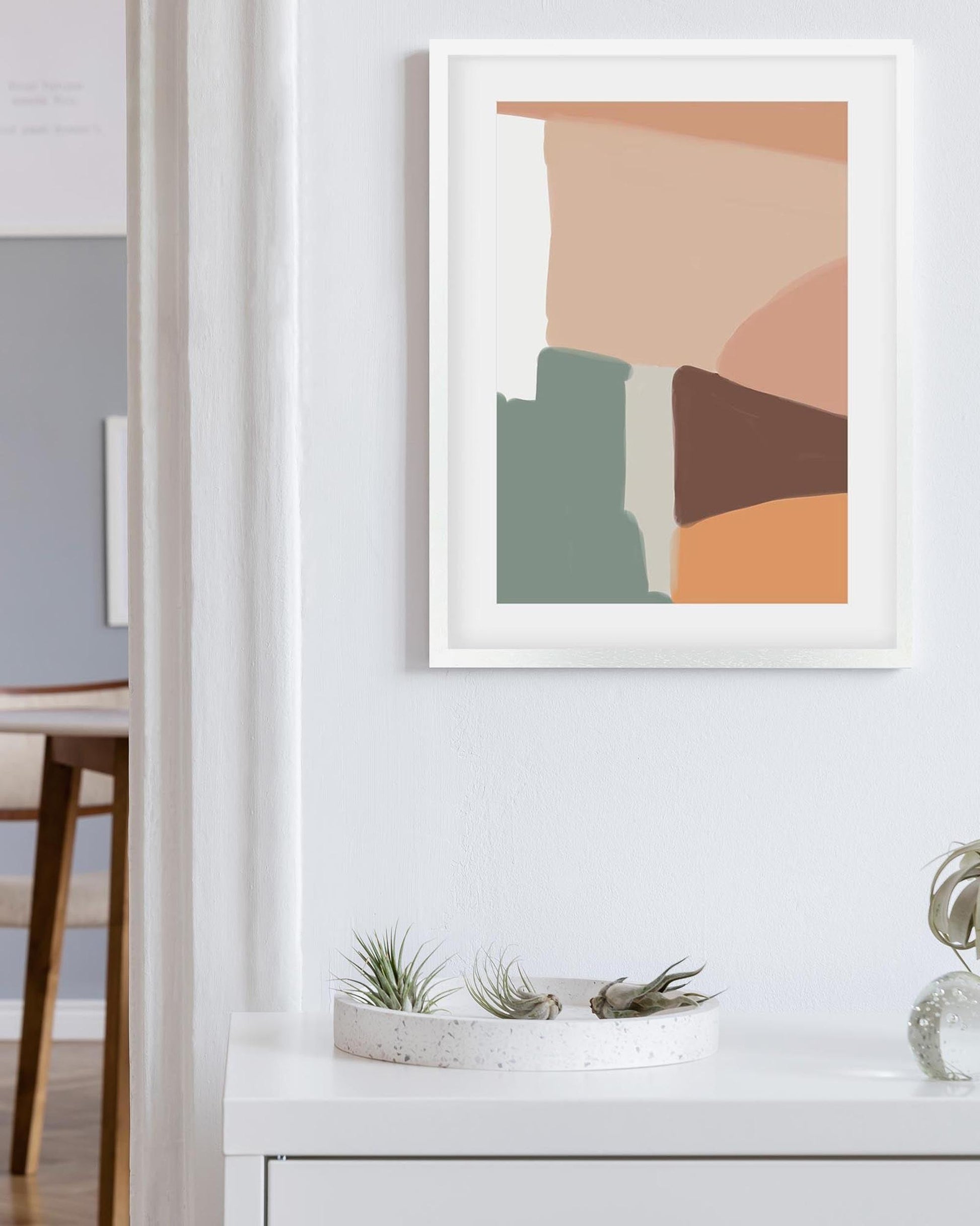 Abstract art print in a white frame on a wall above a white surface with plants.
