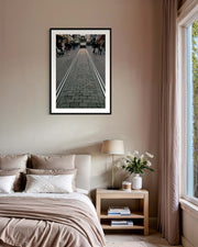 Framed artwork above a bed in a bedroom with a lamp and plant on a nightstand.
