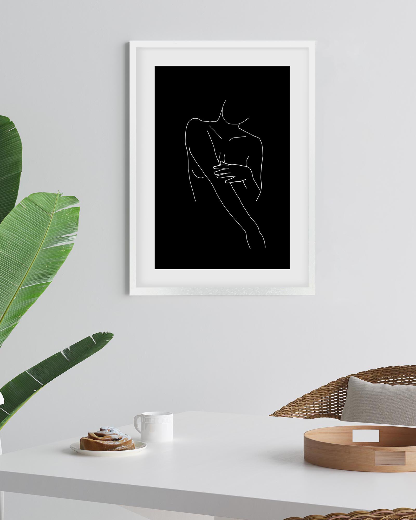 Framed line art of a couple embracing on a black background, placed on a white wall above a table with decor items.