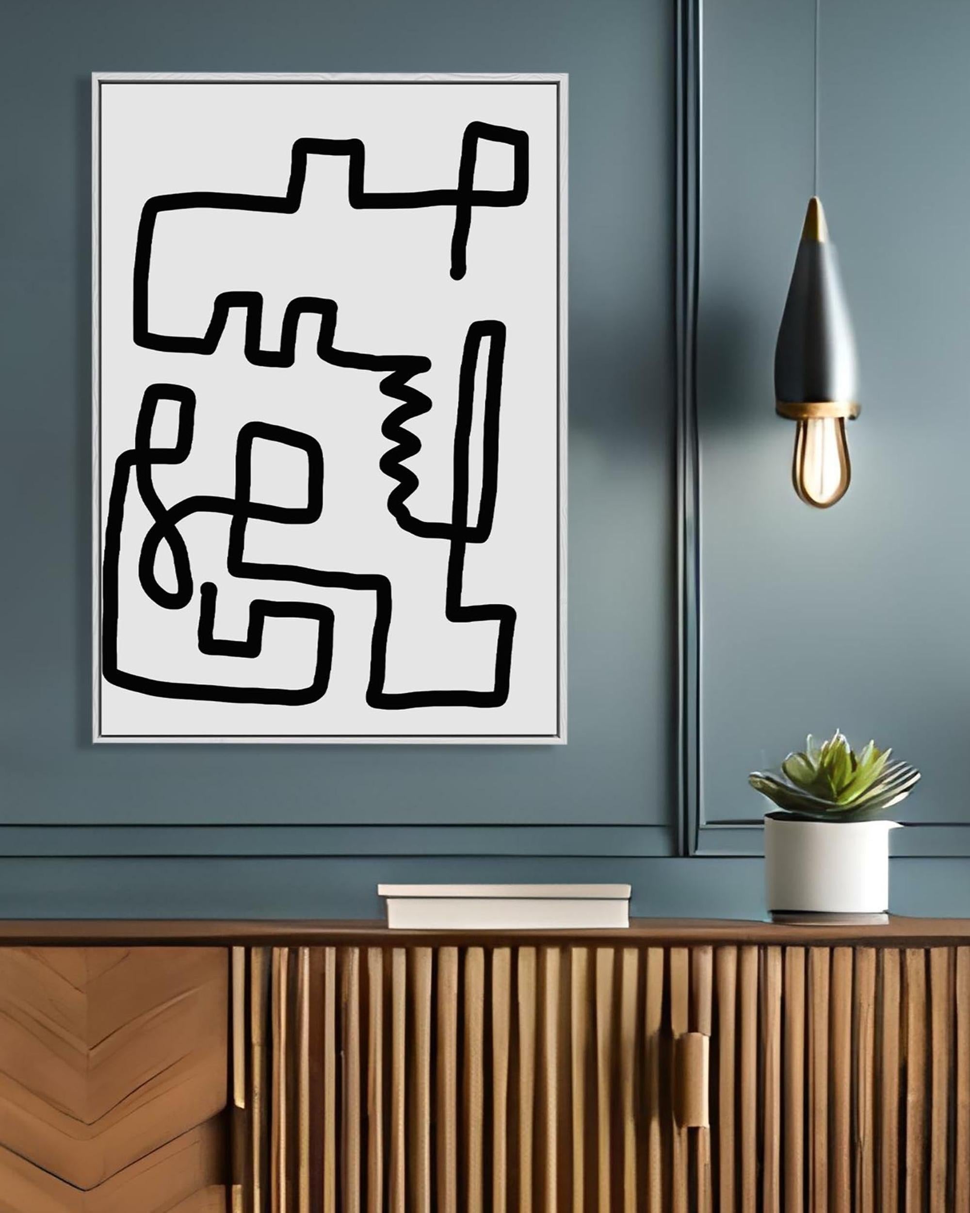A framed canvas with a black and white line art design displayed on a wall above a wooden cabinet, with a small potted plant and a hanging light fixture to the right.