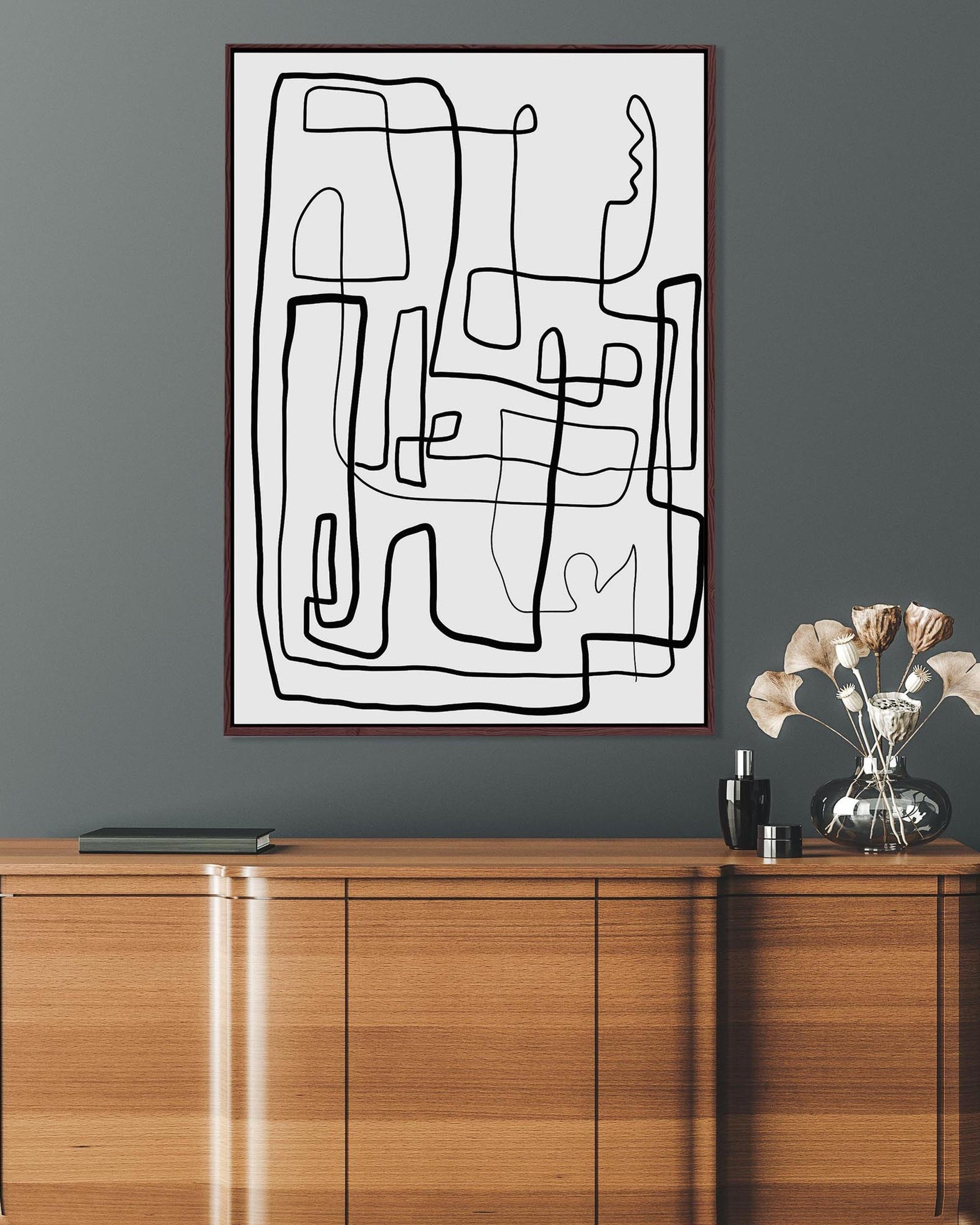 A black and white line art canvas painting depicting a labyrinth, framed and displayed above a wooden cabinet.