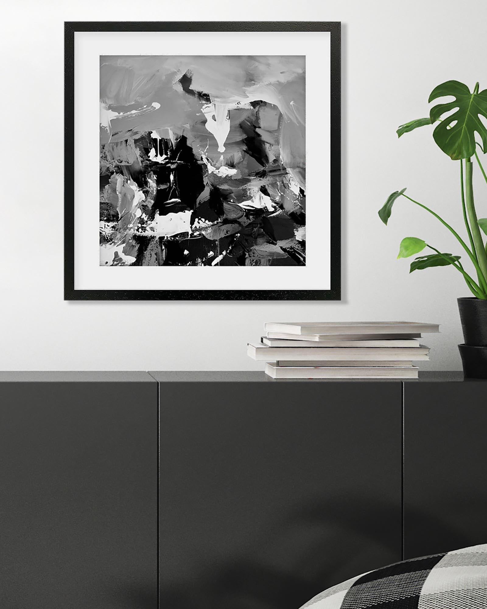 Framed abstract black and white artwork on a wall above a black cabinet with books and a plant.