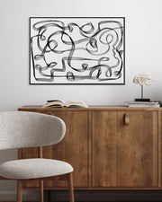 A framed canvas with a geometric black and white line design displayed above a wooden cabinet and a modern chair.