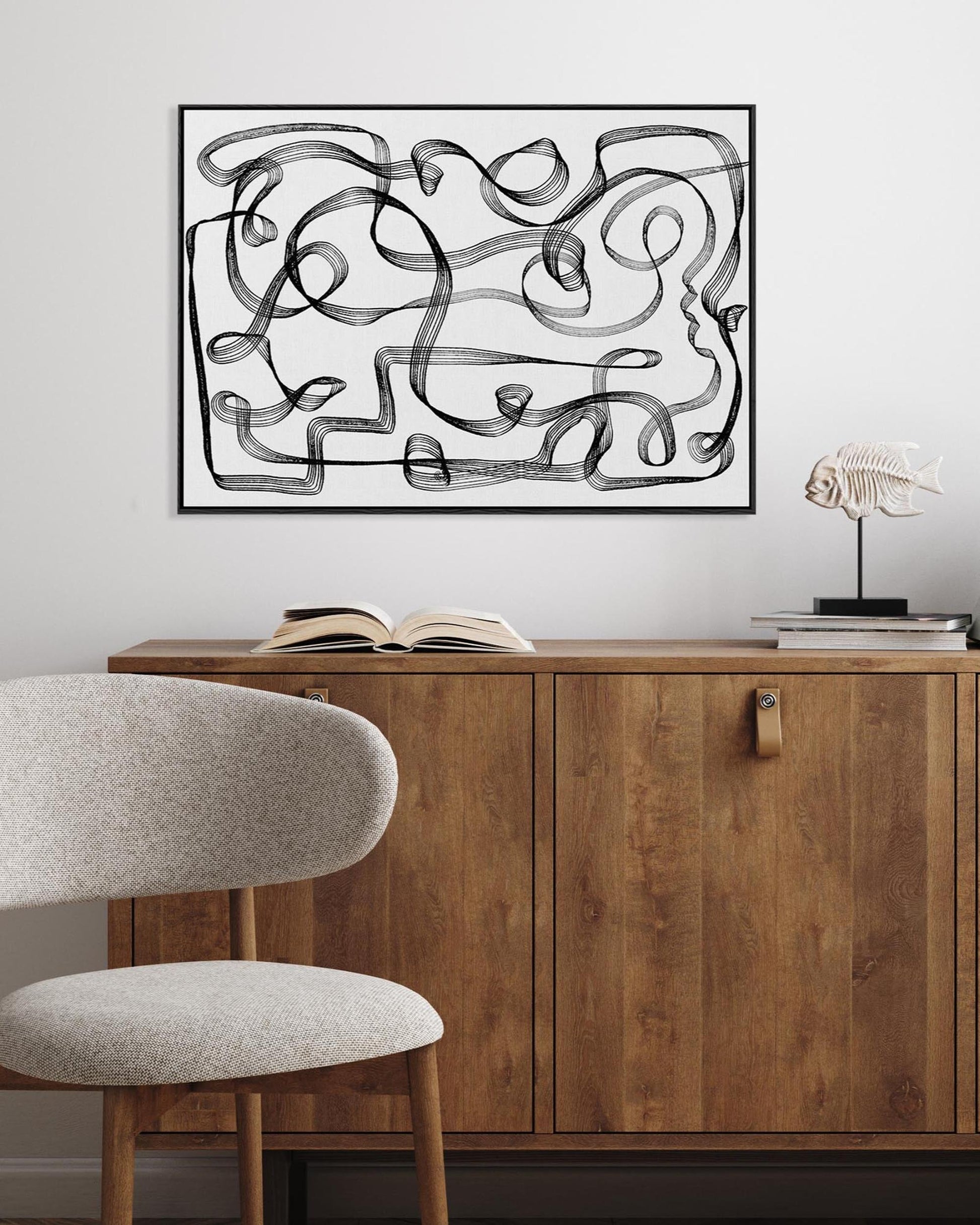 A framed canvas with a geometric black and white line design displayed above a wooden cabinet and a modern chair.