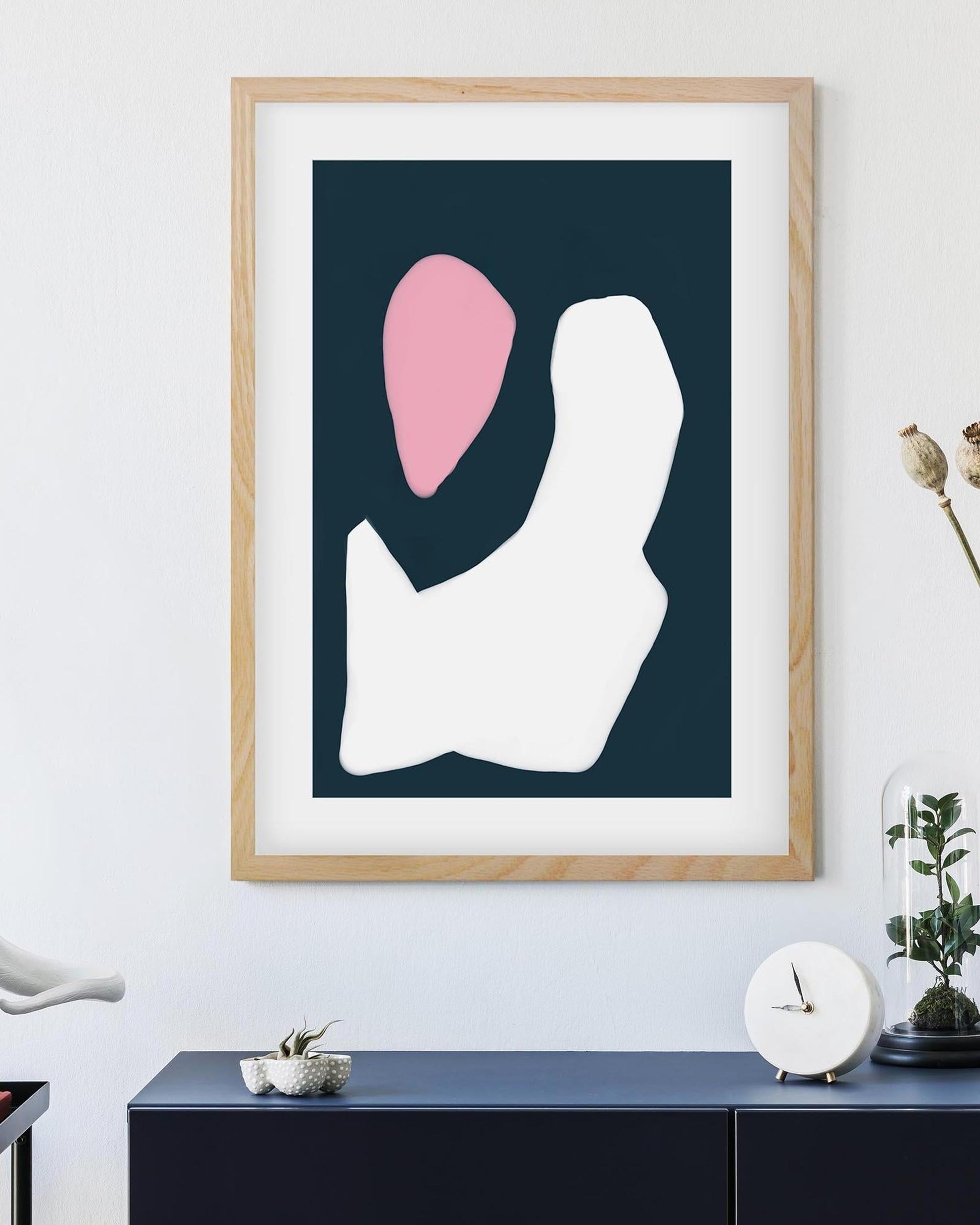 Framed abstract art print with a pink and white shape on a dark blue background, displayed in a room.