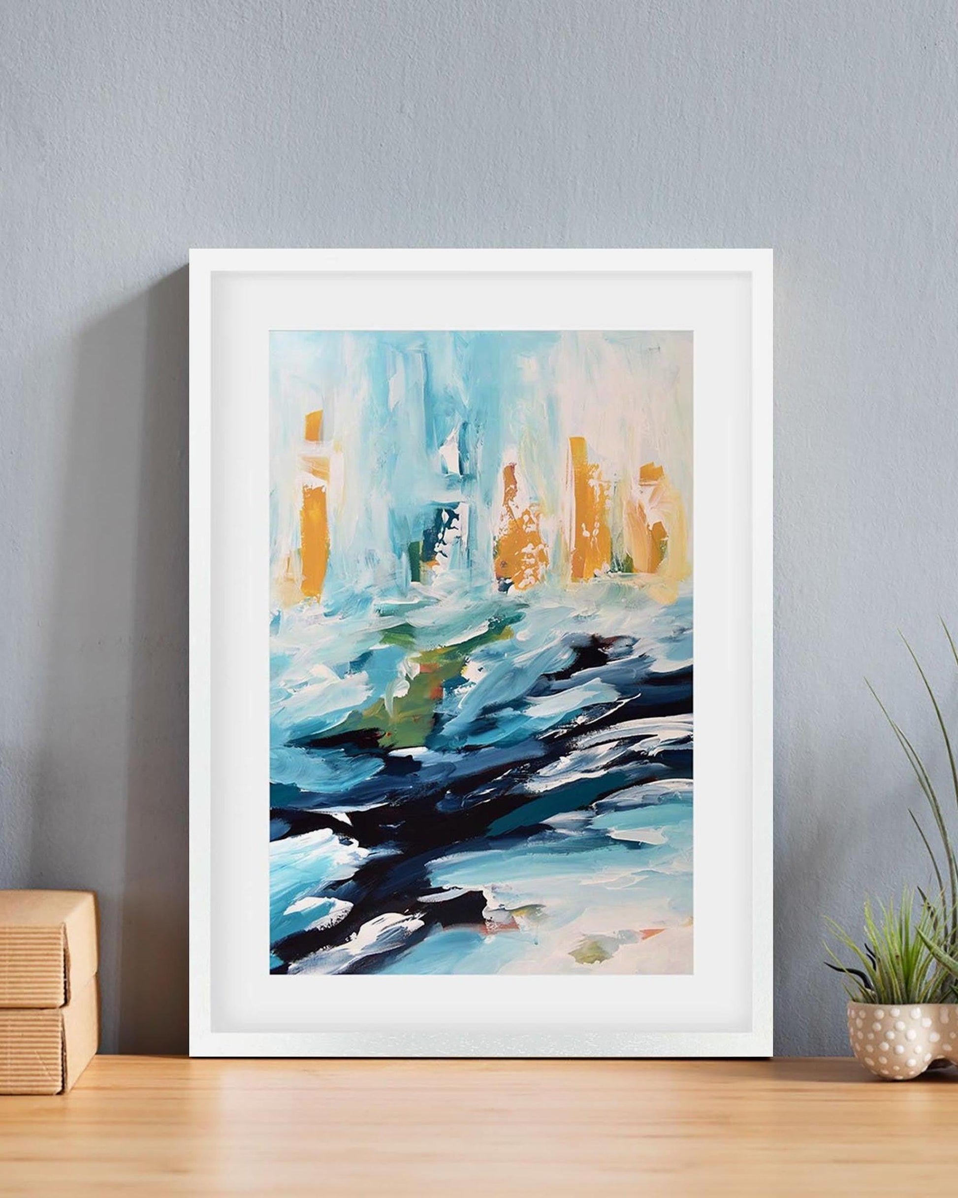 Framed abstract painting on a wall with a plant and wooden box below