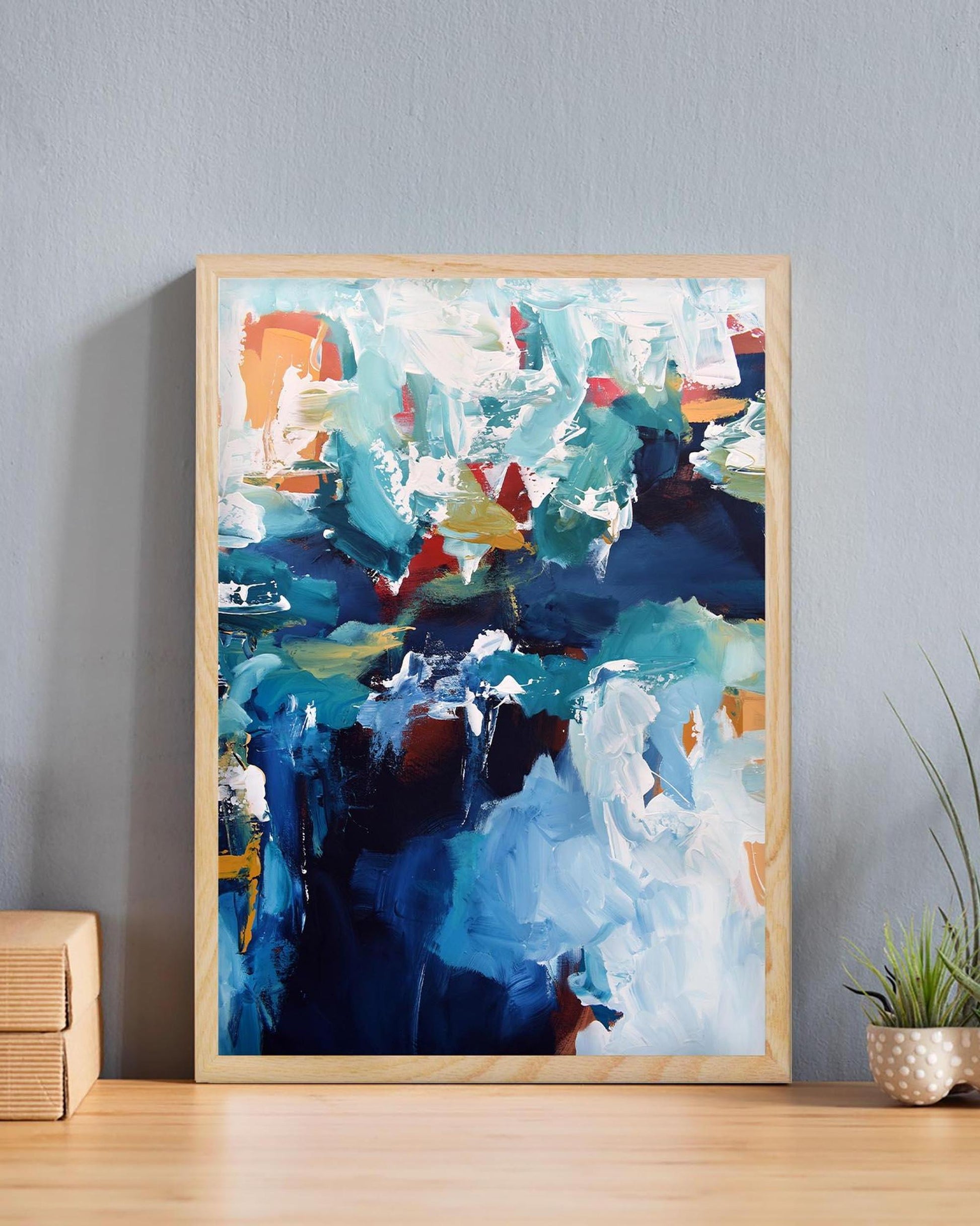 Abstract painting in a wooden frame on a wall with a plant and wooden side table.