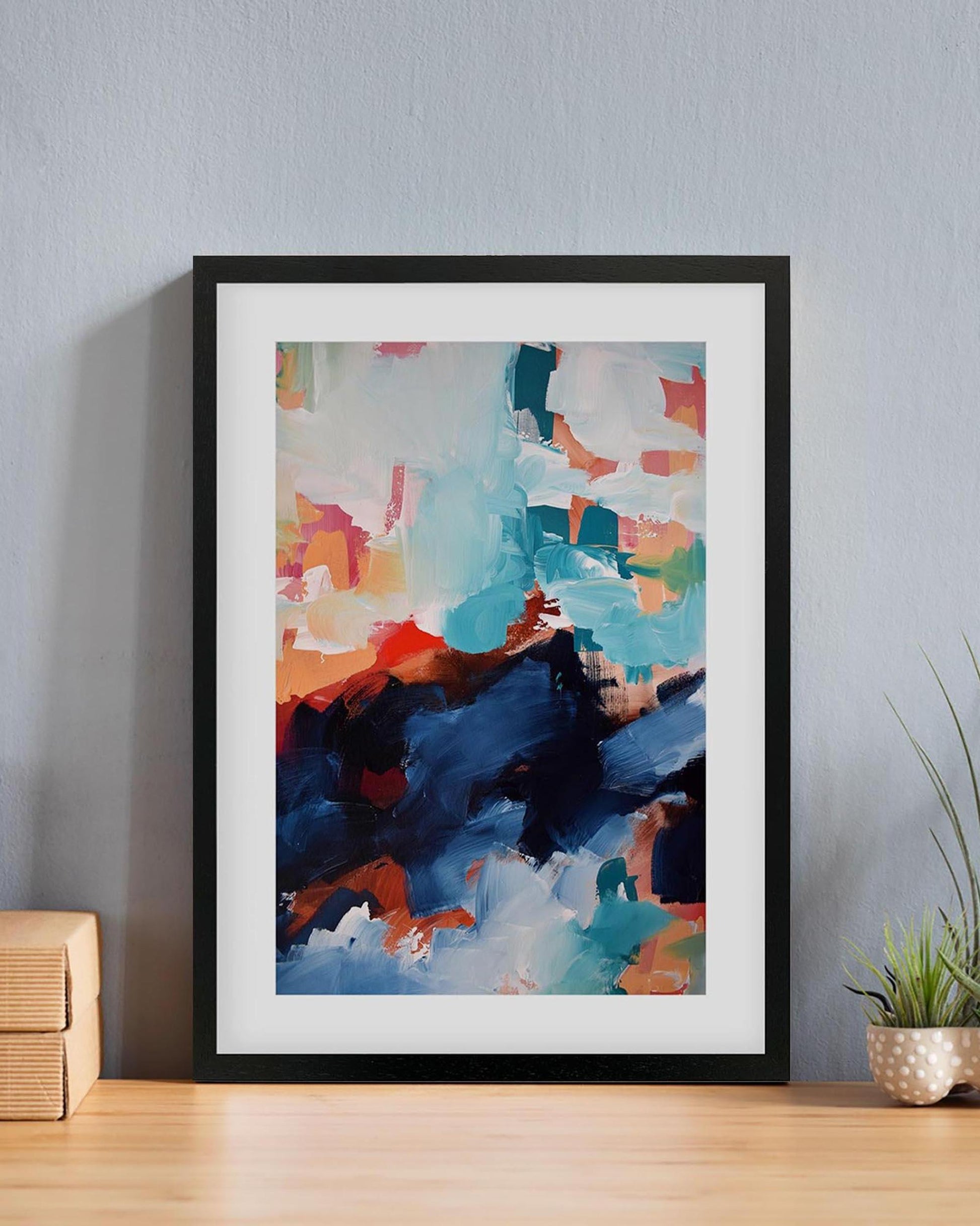 Framed abstract art print on a wall with a plant and wooden box in the background