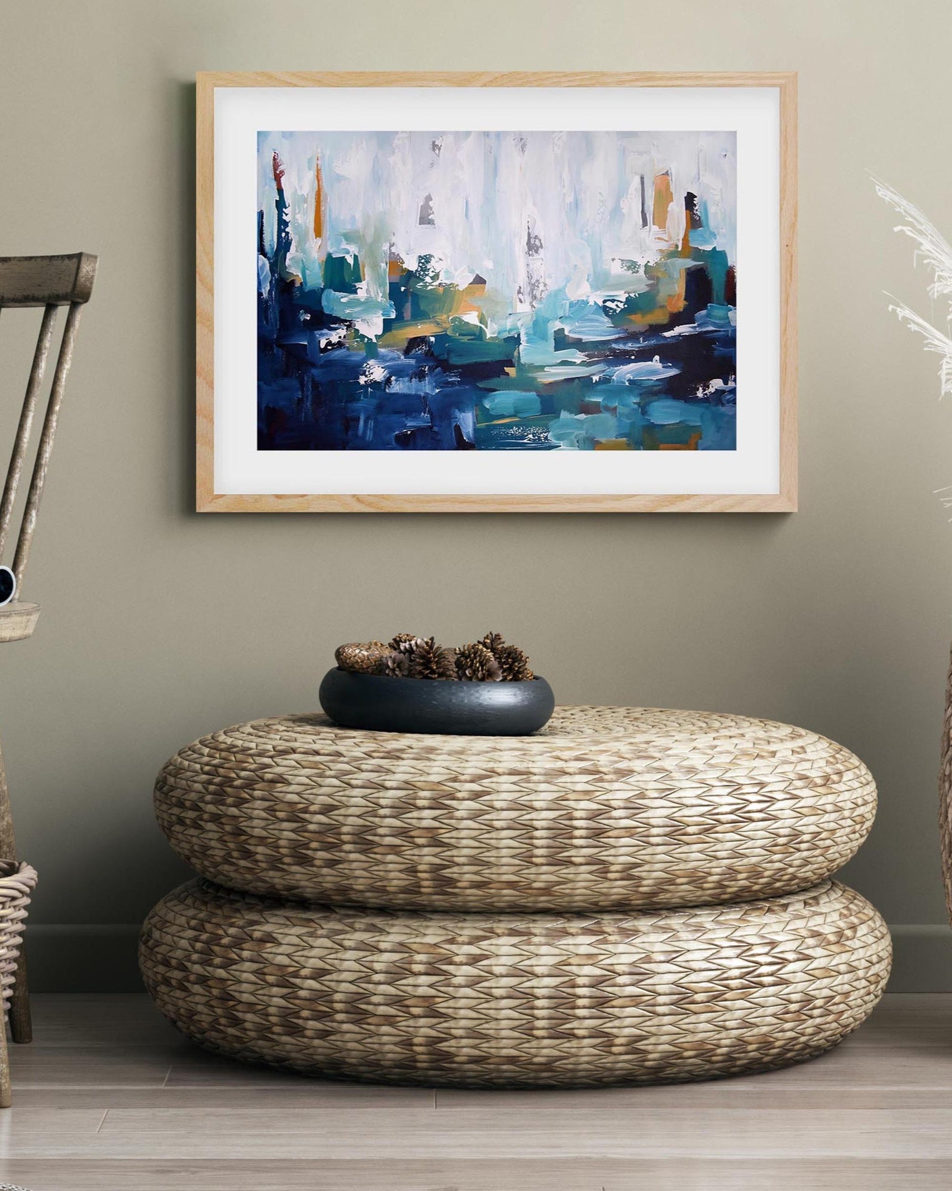 Abstract painting on a wall above a woven ottoman with a decorative bowl.