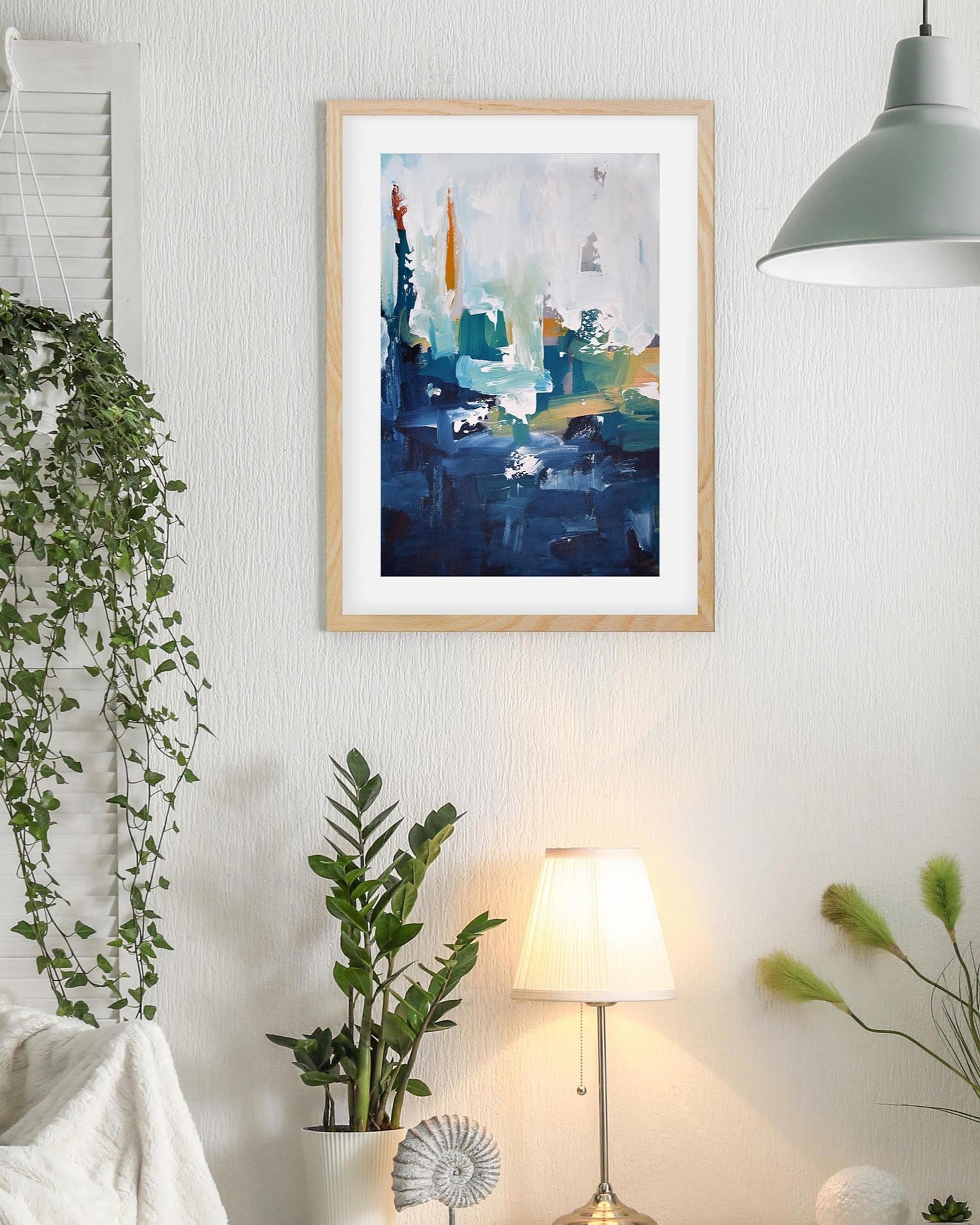 Framed abstract art piece on a wall with plants and a lamp in the room.