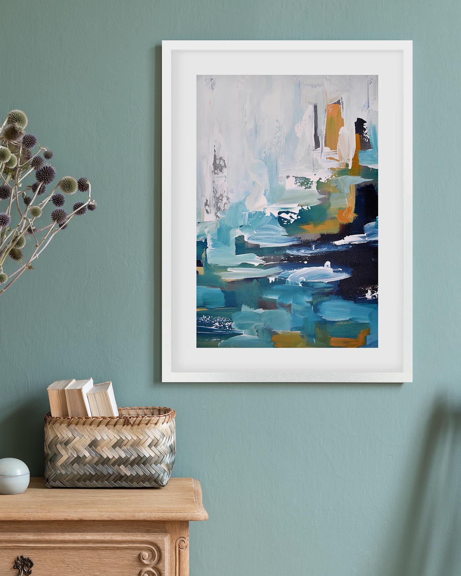 Framed abstract art piece on a teal wall with a wooden cabinet and basket below.