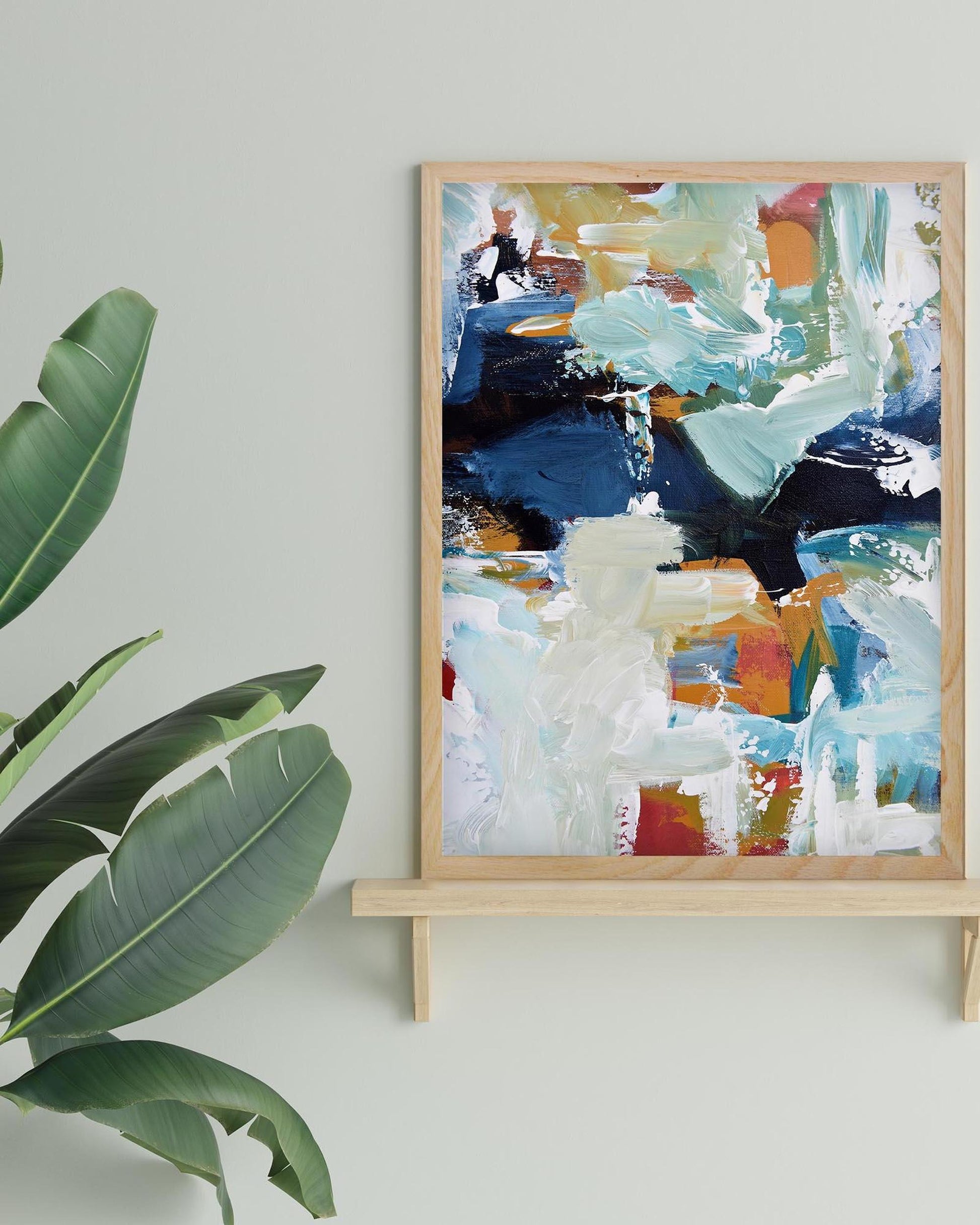 Abstract painting in a wooden frame on a white wall with a plant to the left.