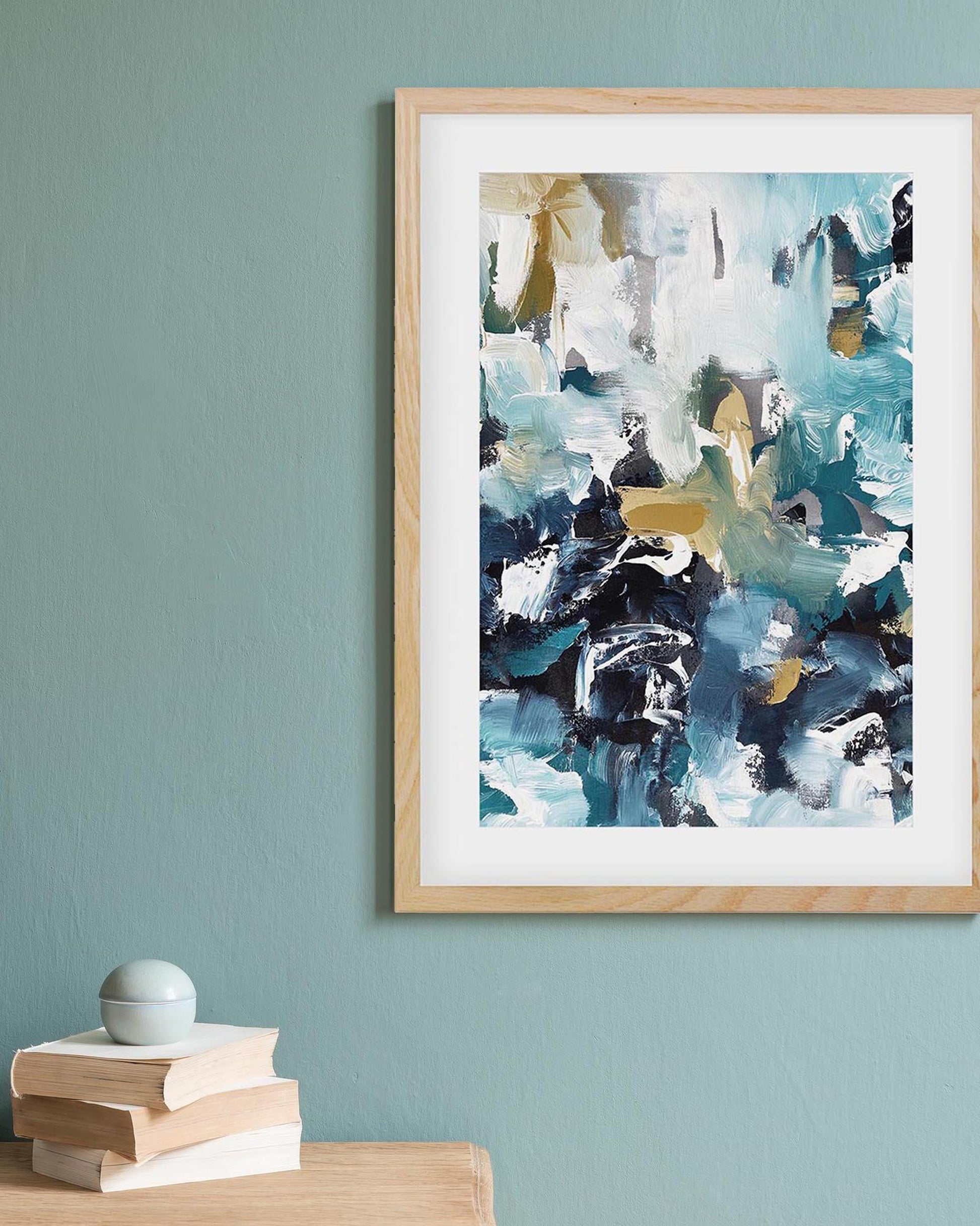 Framed abstract art piece on a light blue wall with books and a spherical object below.