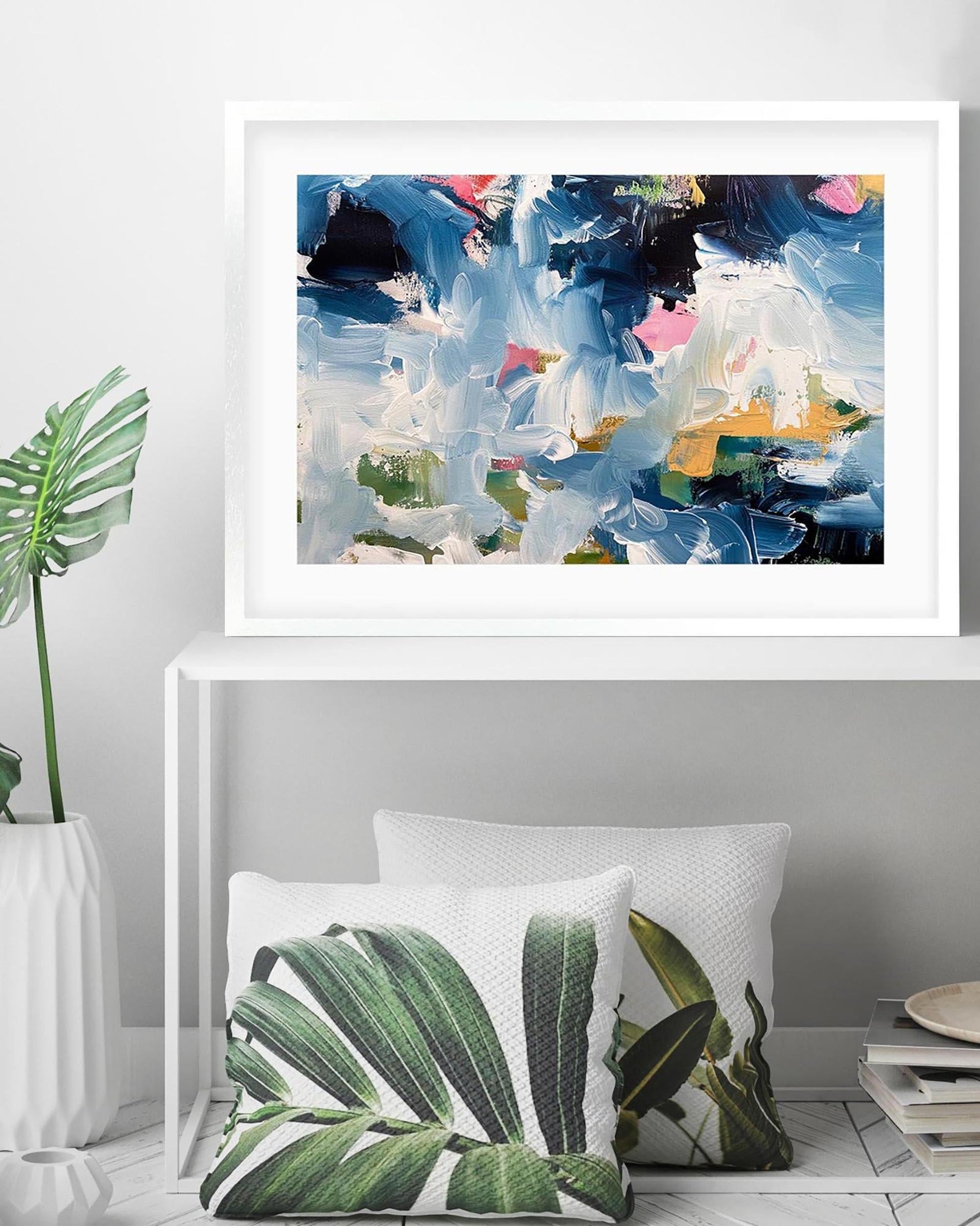 Abstract painting in a white frame above two leaf-patterned cushions on a shelf.