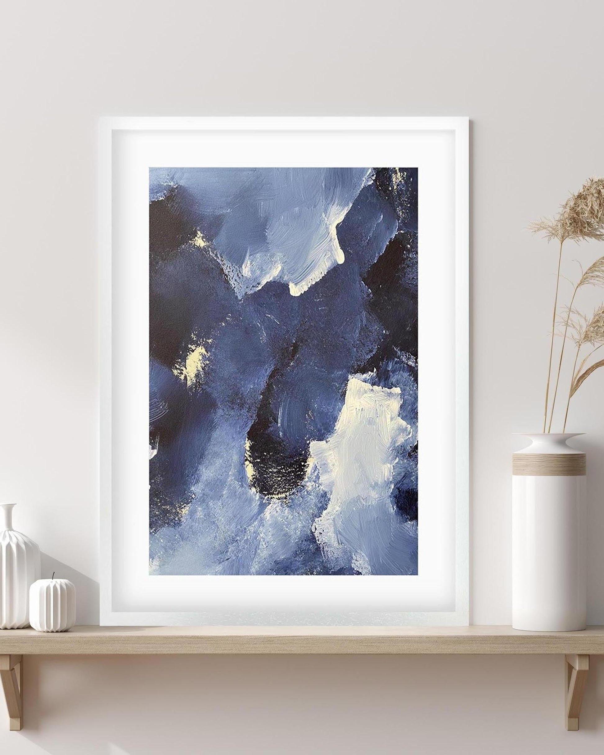 Framed abstract art print with blue and white colors on a wall above a wooden shelf.