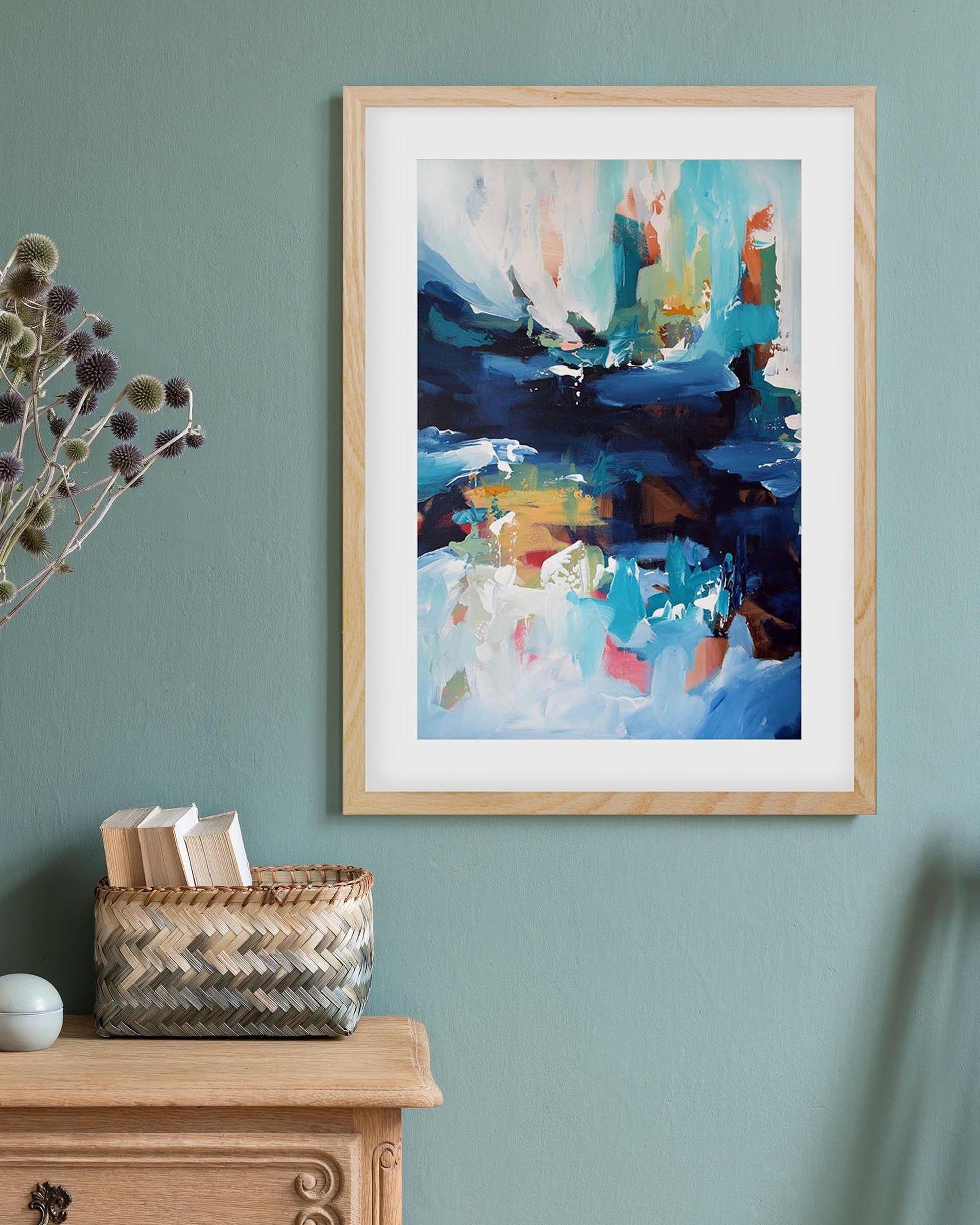 Framed abstract painting on a teal wall with a wooden cabinet and basket below.