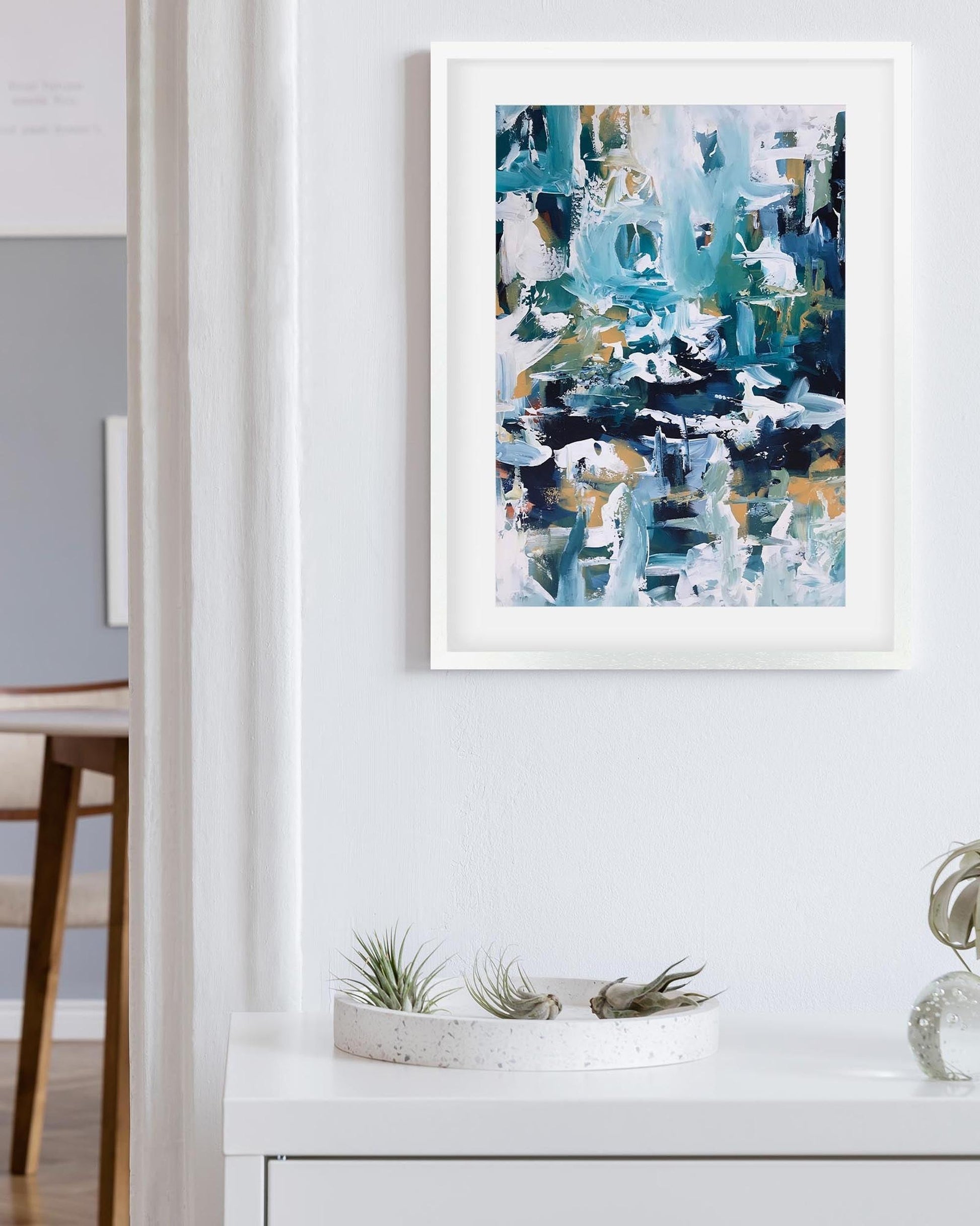Abstract art print in a white frame on a wall with a dining area in the background