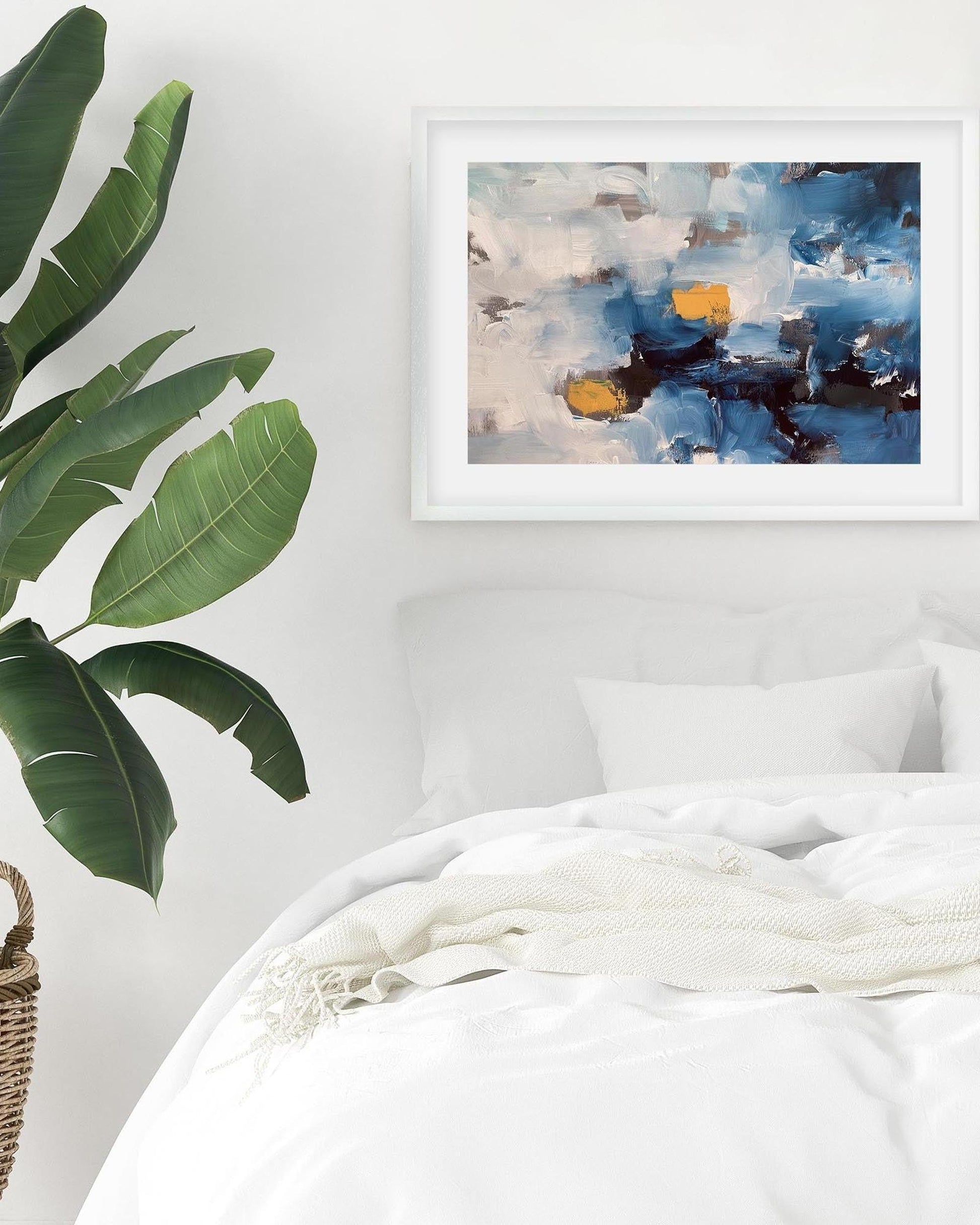 Framed abstract painting on a wall above a bed with a plant to the left.
