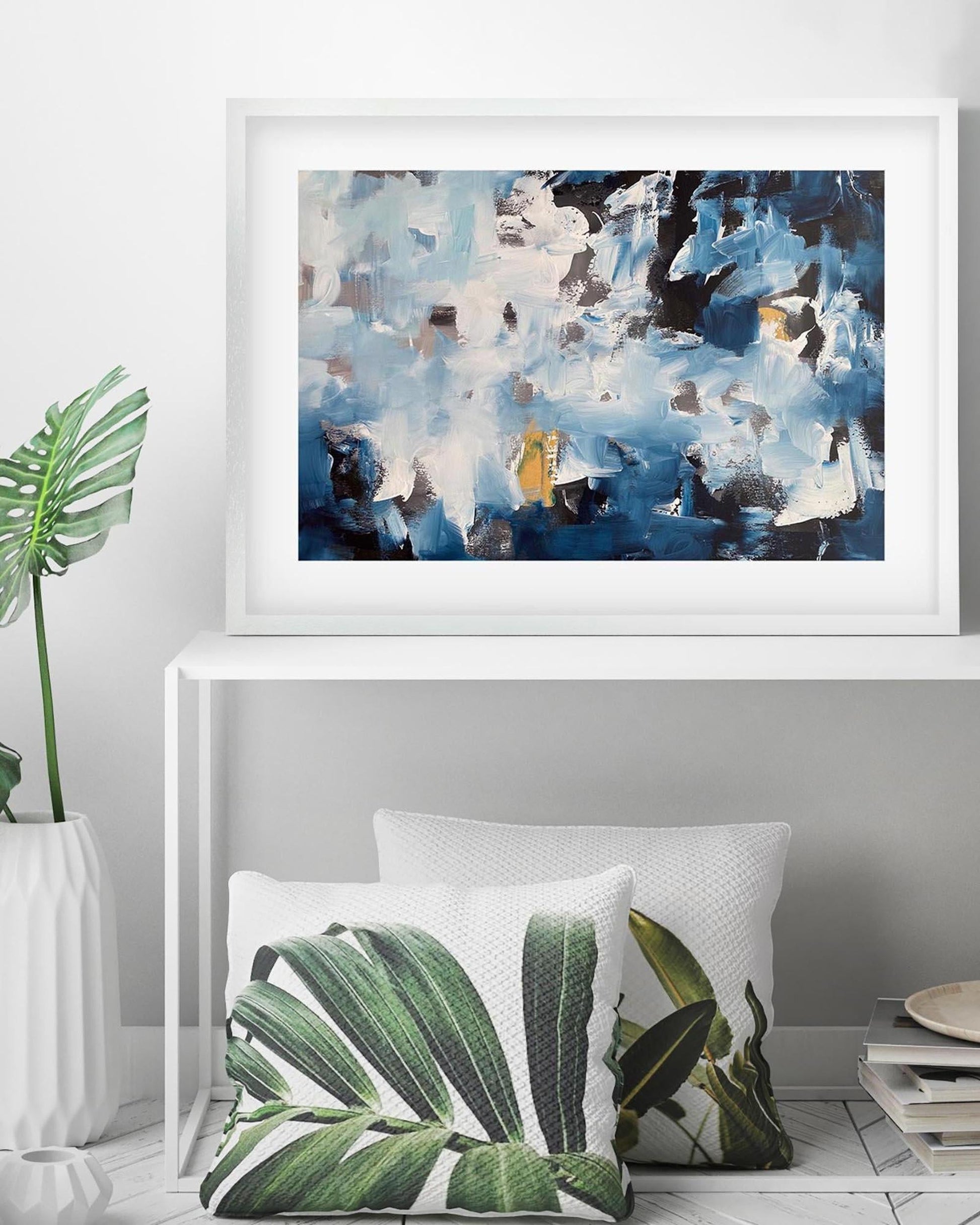 Framed abstract art above two leaf-patterned cushions on a white surface.