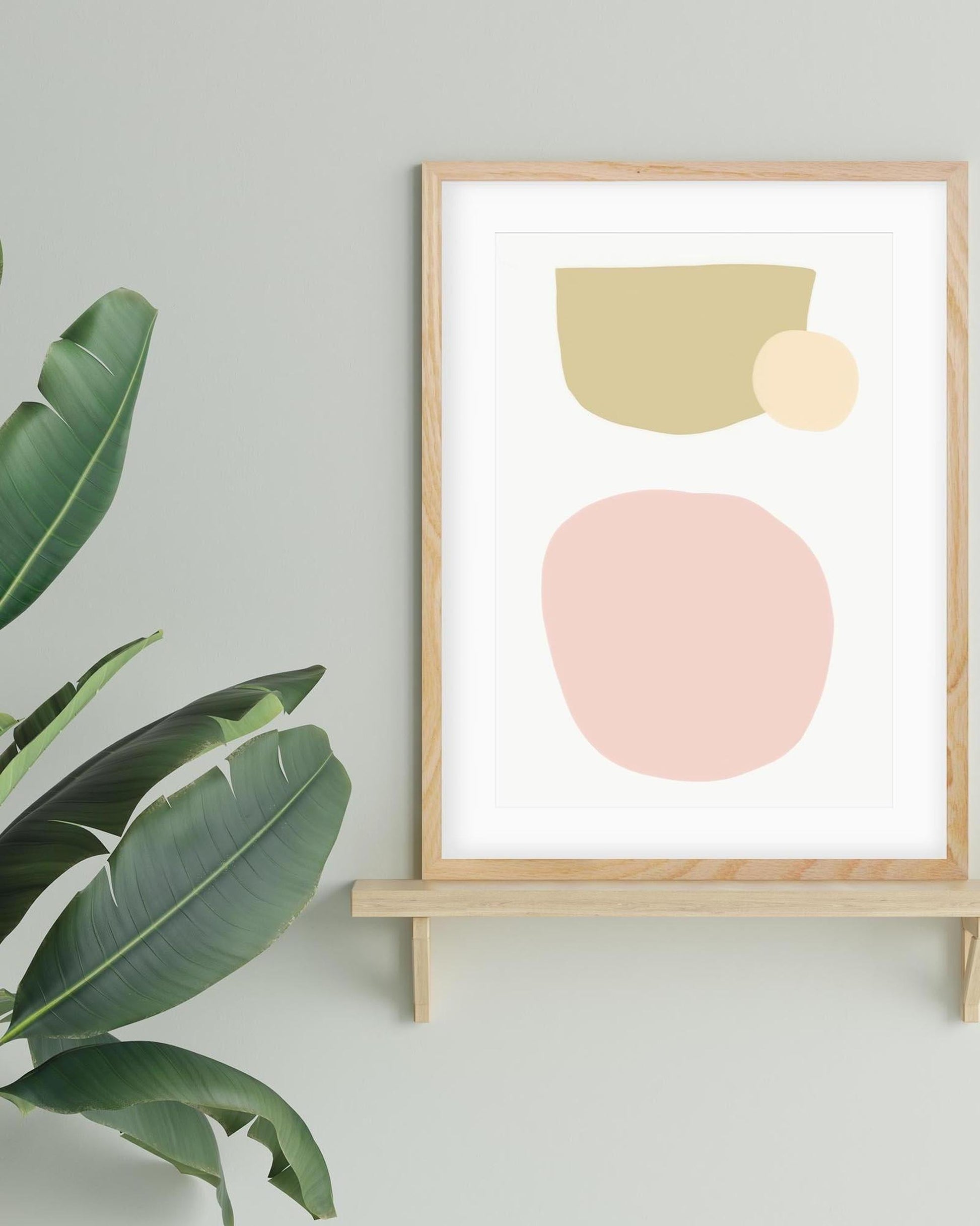 Framed abstract art print with geometric shapes on a stand against a light gray wall, next to a plant.