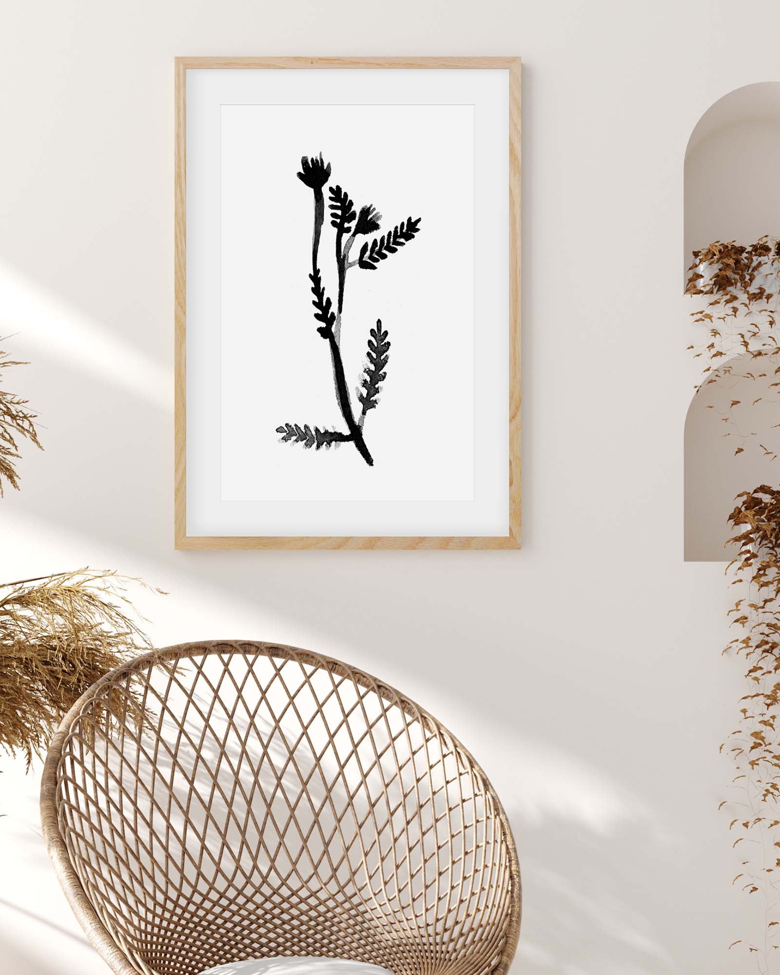 Framed black botanical print on a white wall with a wicker chair and decorative elements.