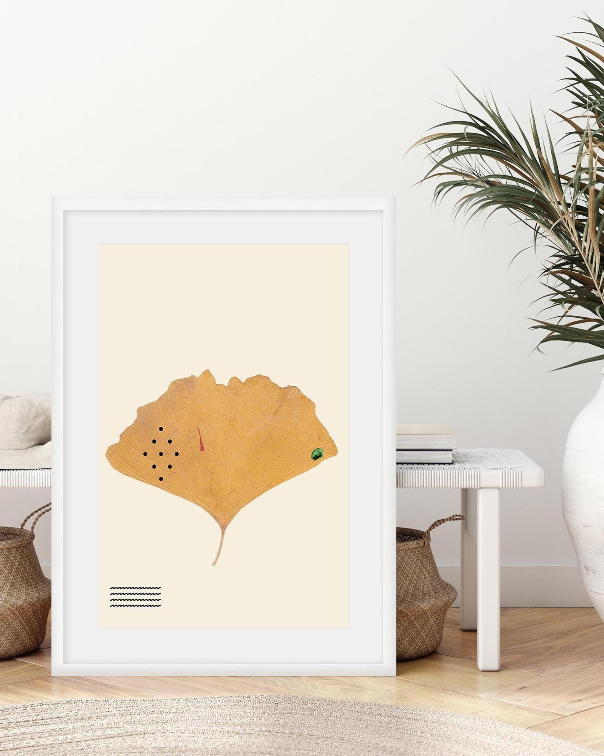 Framed artwork of a yellow leaf on a beige background in a room with a plant and table.