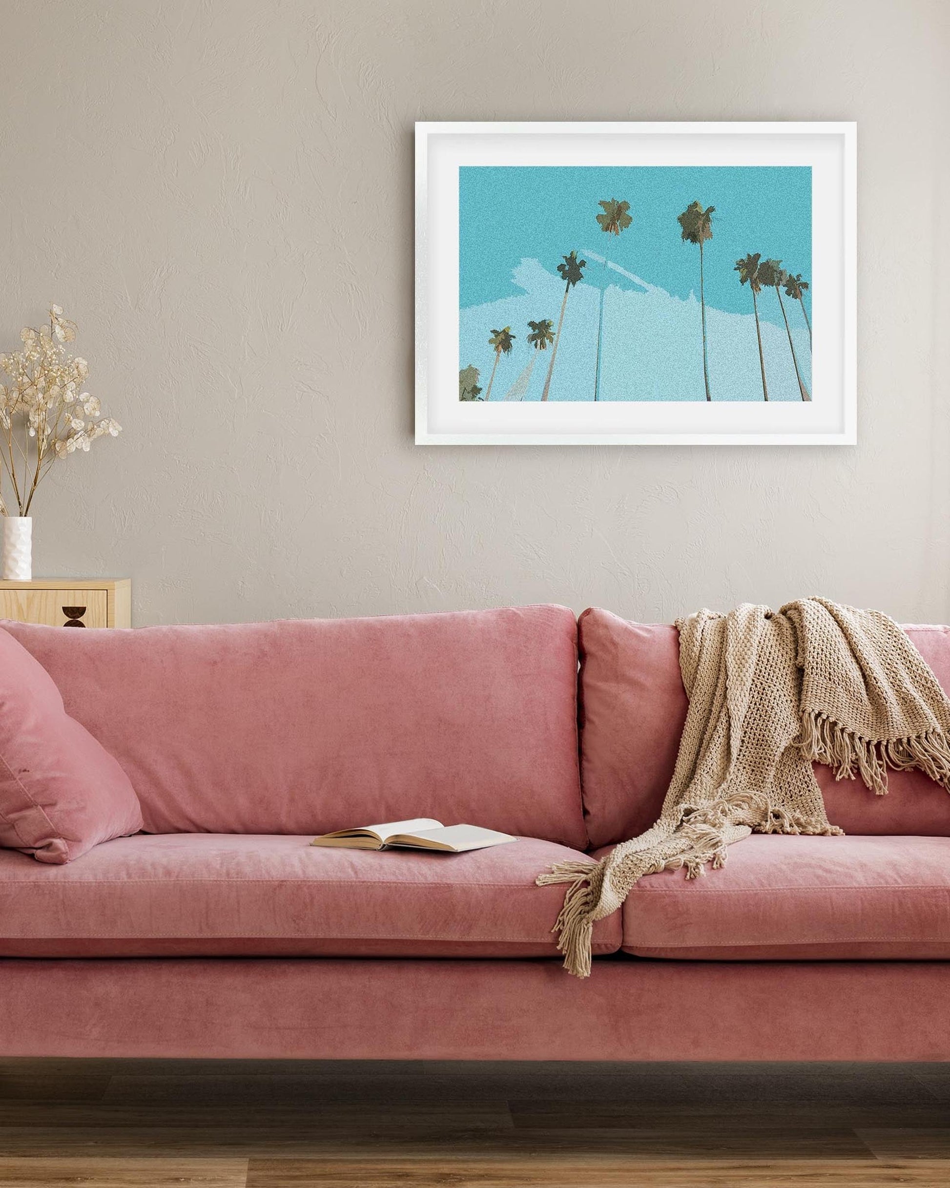 Pink sofa in a living room with a framed artwork of palm trees on the wall.