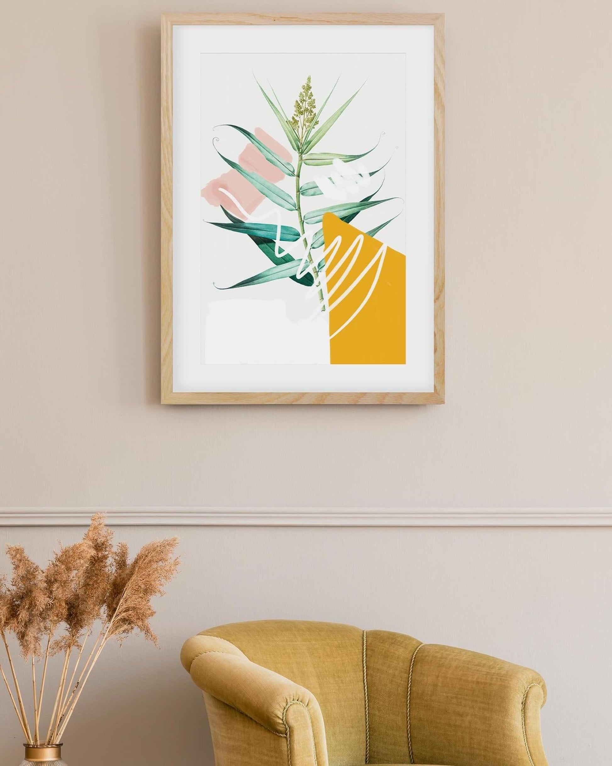 Framed abstract art print with green leaves and geometric shapes on a beige wall above a mustard yellow armchair.