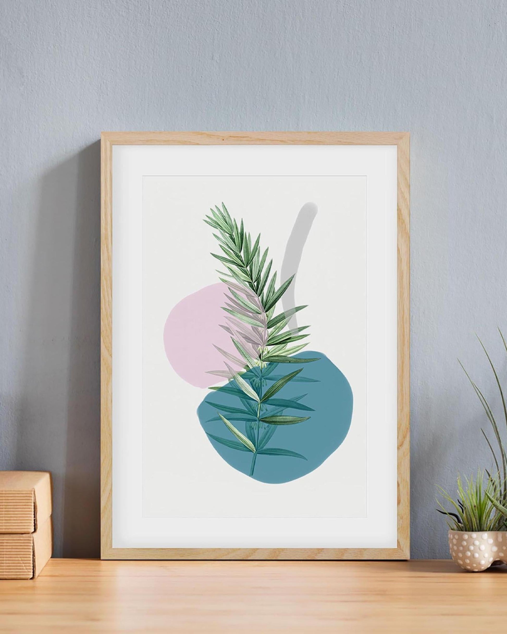 Framed artwork with green leaves on a white background, placed on a wooden surface against a gray wall.