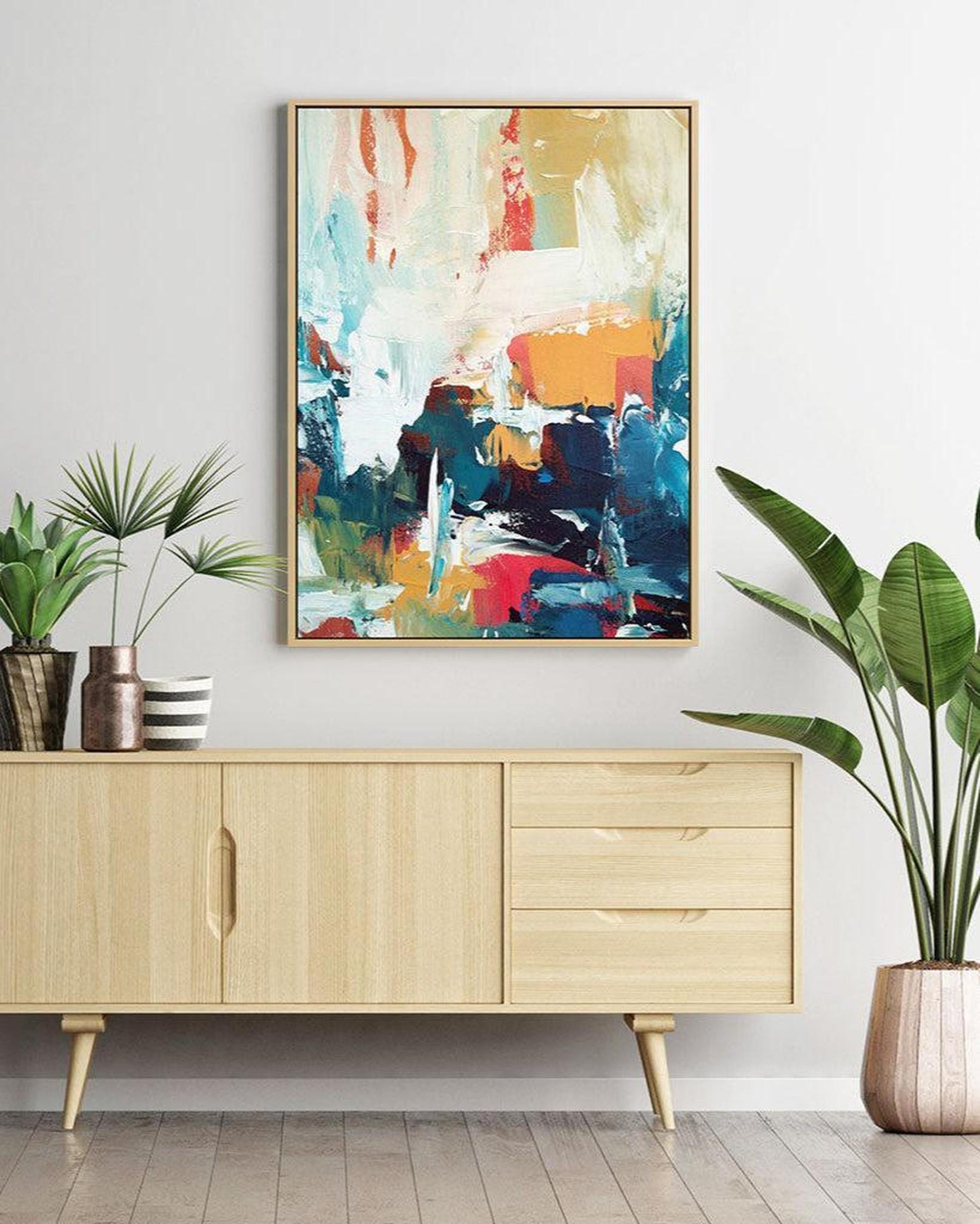 Colorful abstract painting on a wall above a wooden cabinet with plants around.