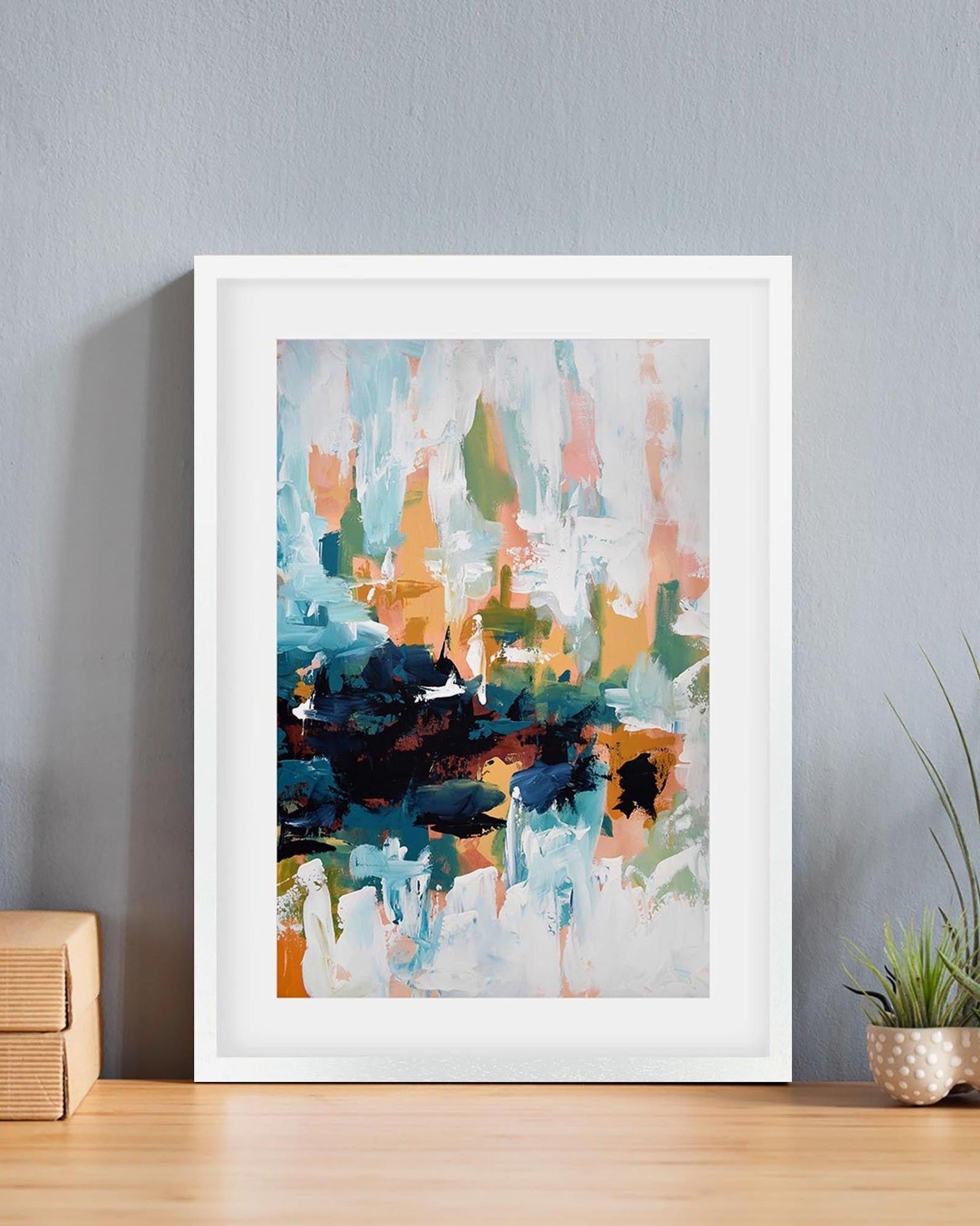 Framed abstract art print on a wall with a plant and wooden box below.