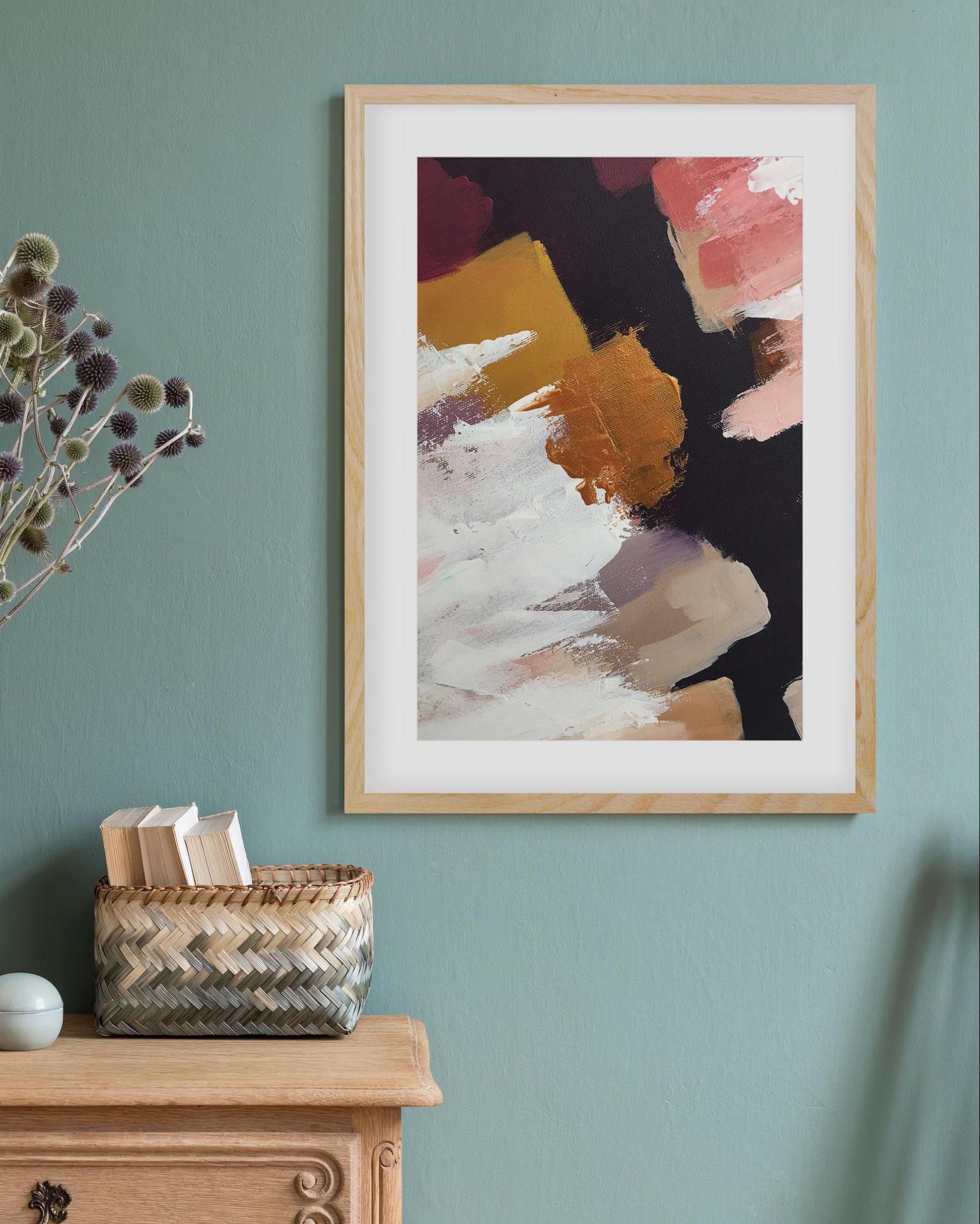 Framed abstract art on a wall with a vase of dried flowers and a basket below.