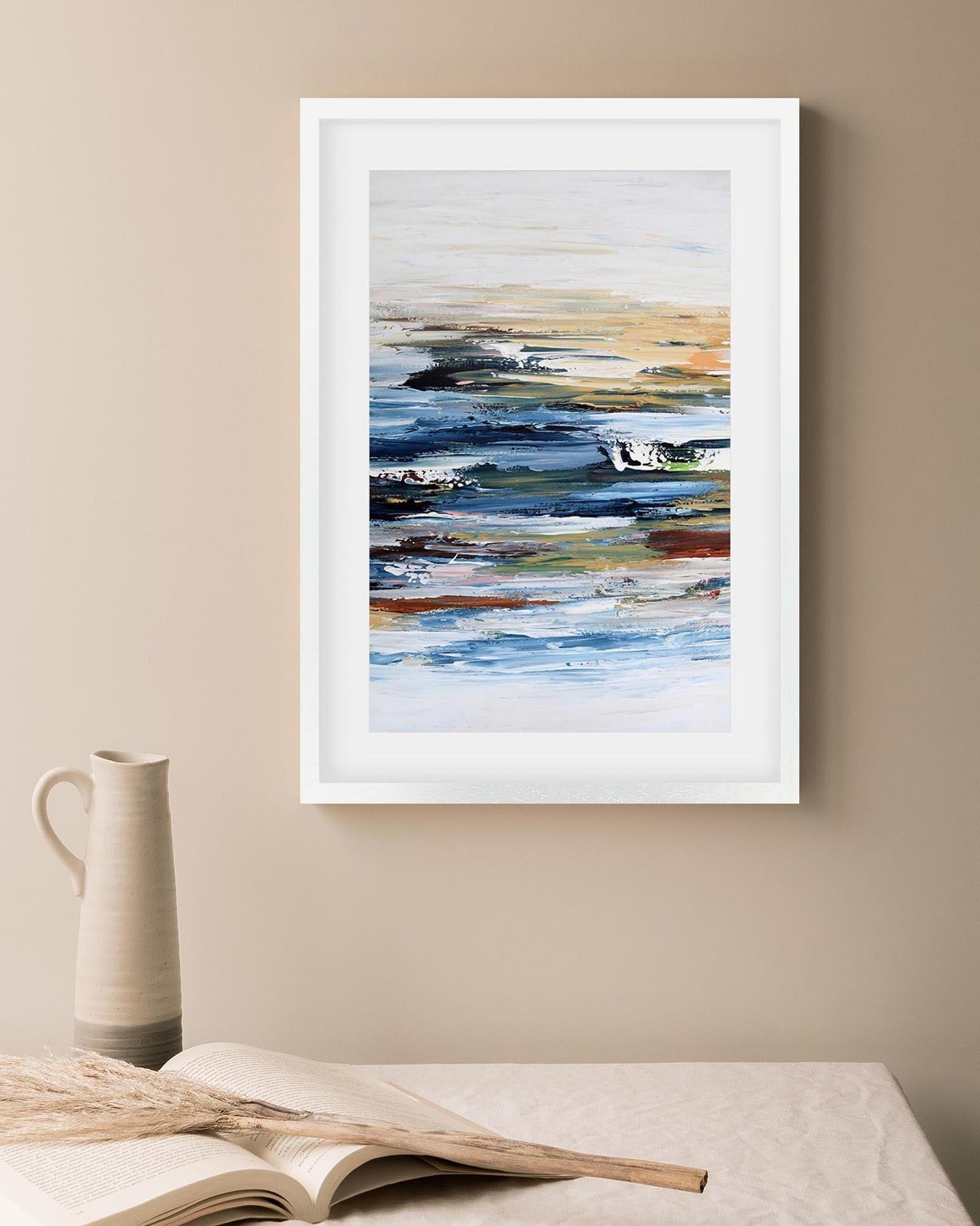 Framed abstract art on a beige wall with a pitcher and book below.