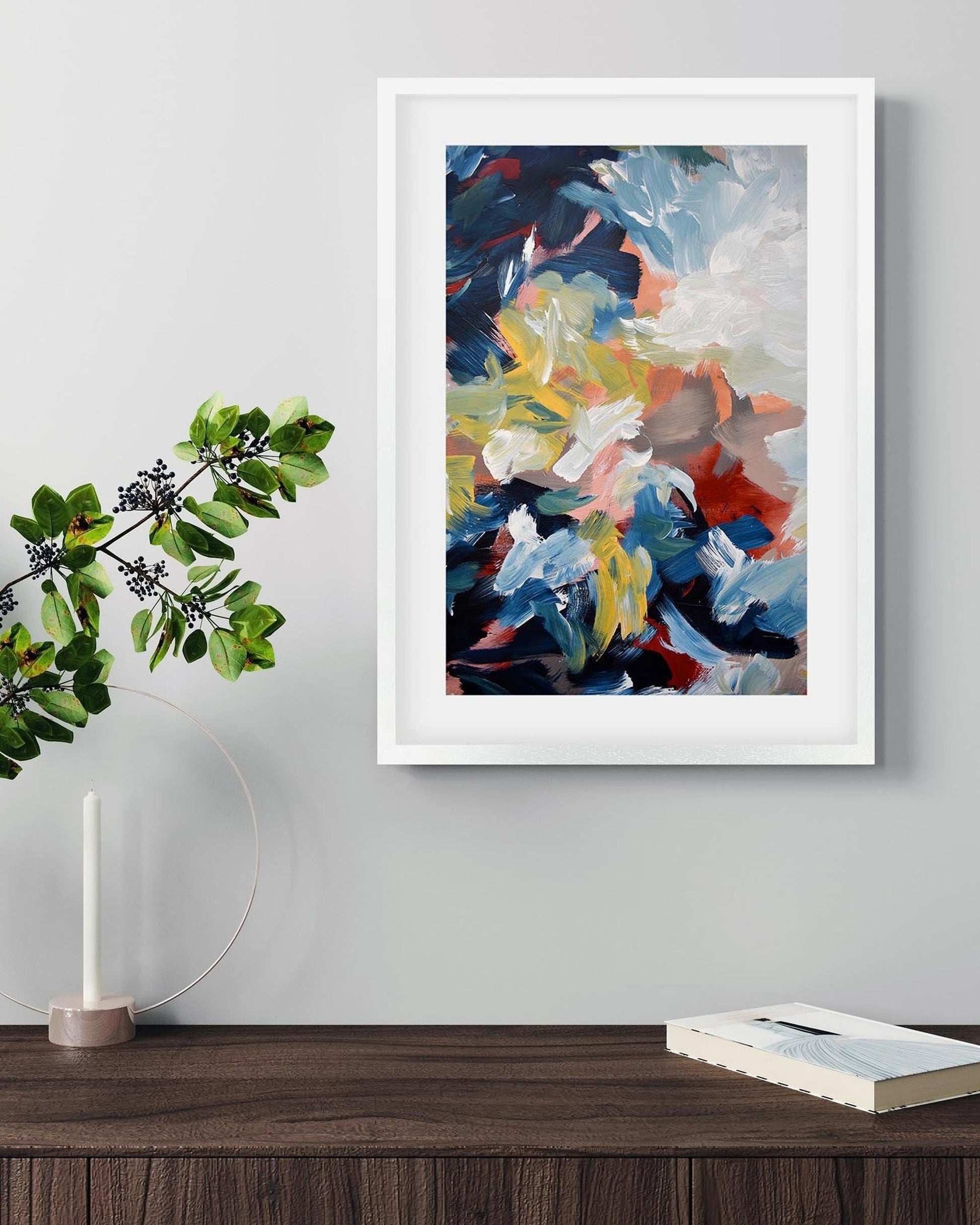 Framed abstract painting on a wall with a plant and book on a wooden surface below.