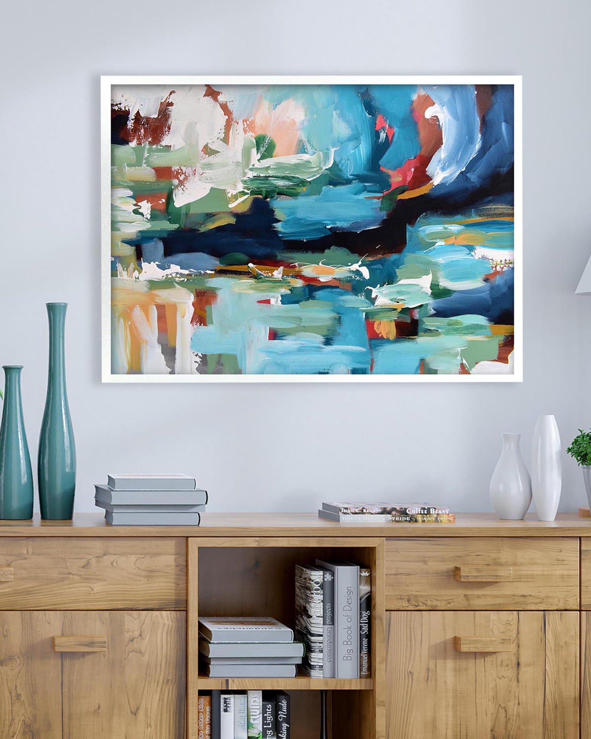 Abstract painting in a white frame on a wall above a wooden cabinet with books and decor items.