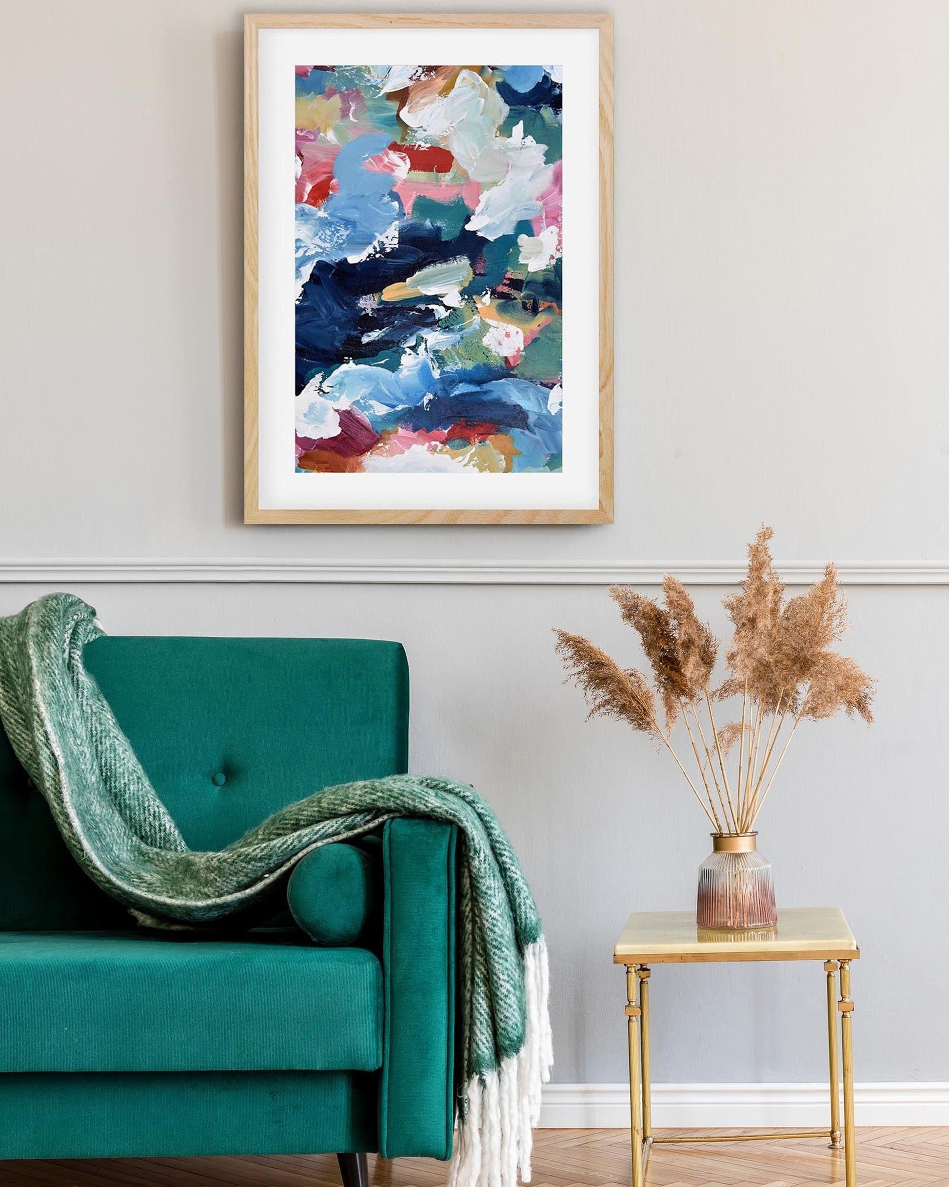 Framed abstract art above a teal chair with a gold side table and decorative plant.