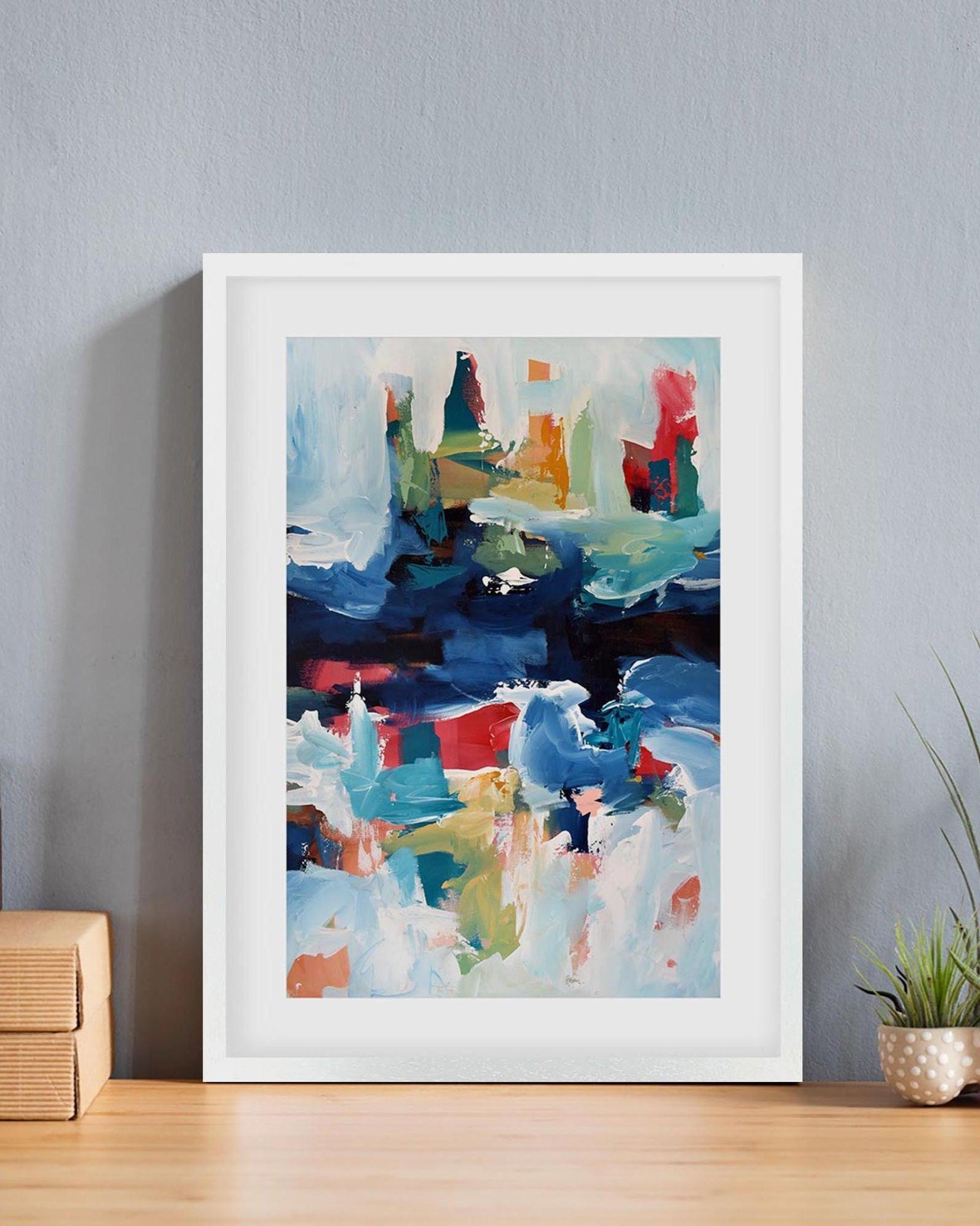 Framed abstract art print on a wall with a plant and wooden box below.