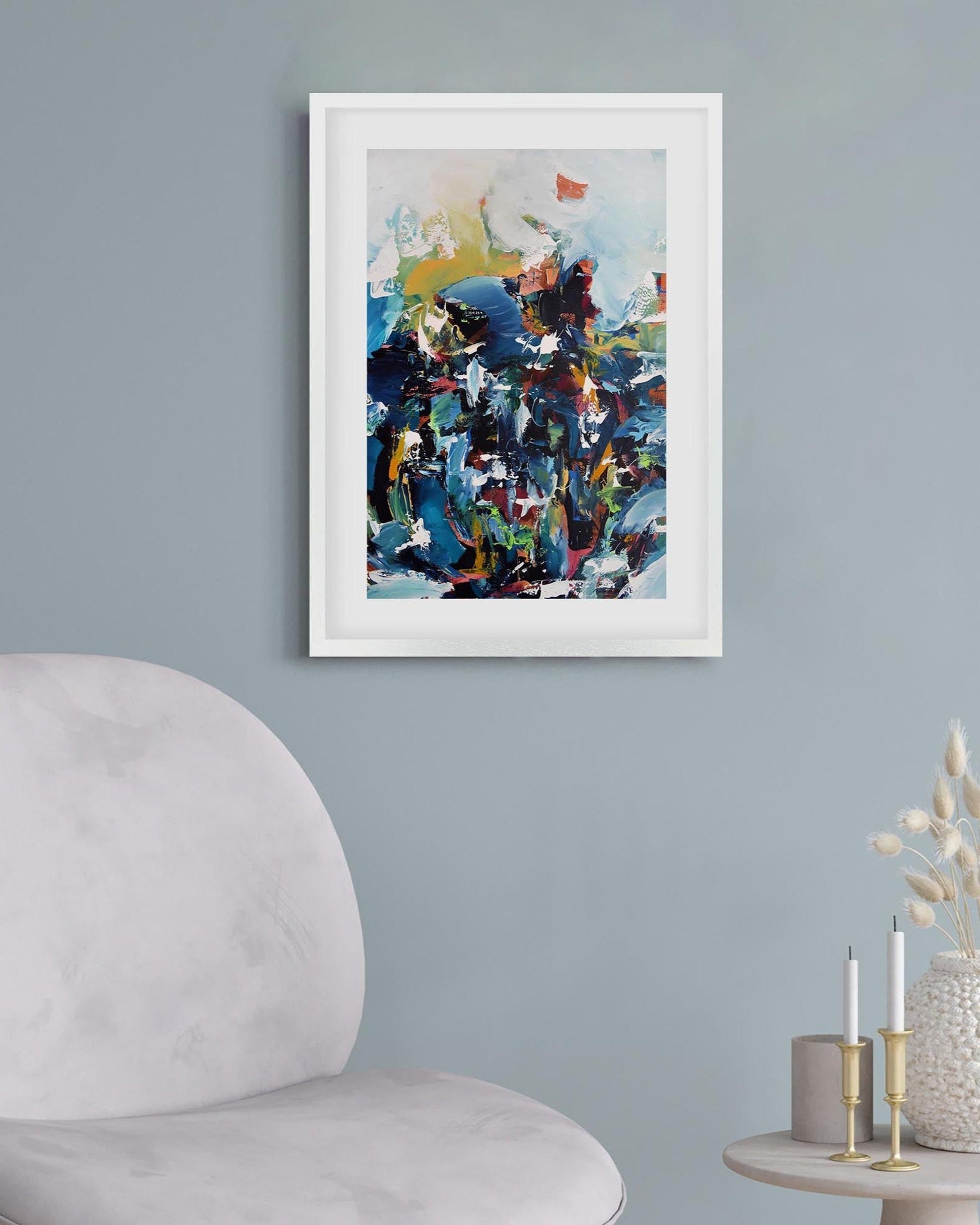 Framed abstract art piece on a light blue wall with a chair and candles in the foreground.