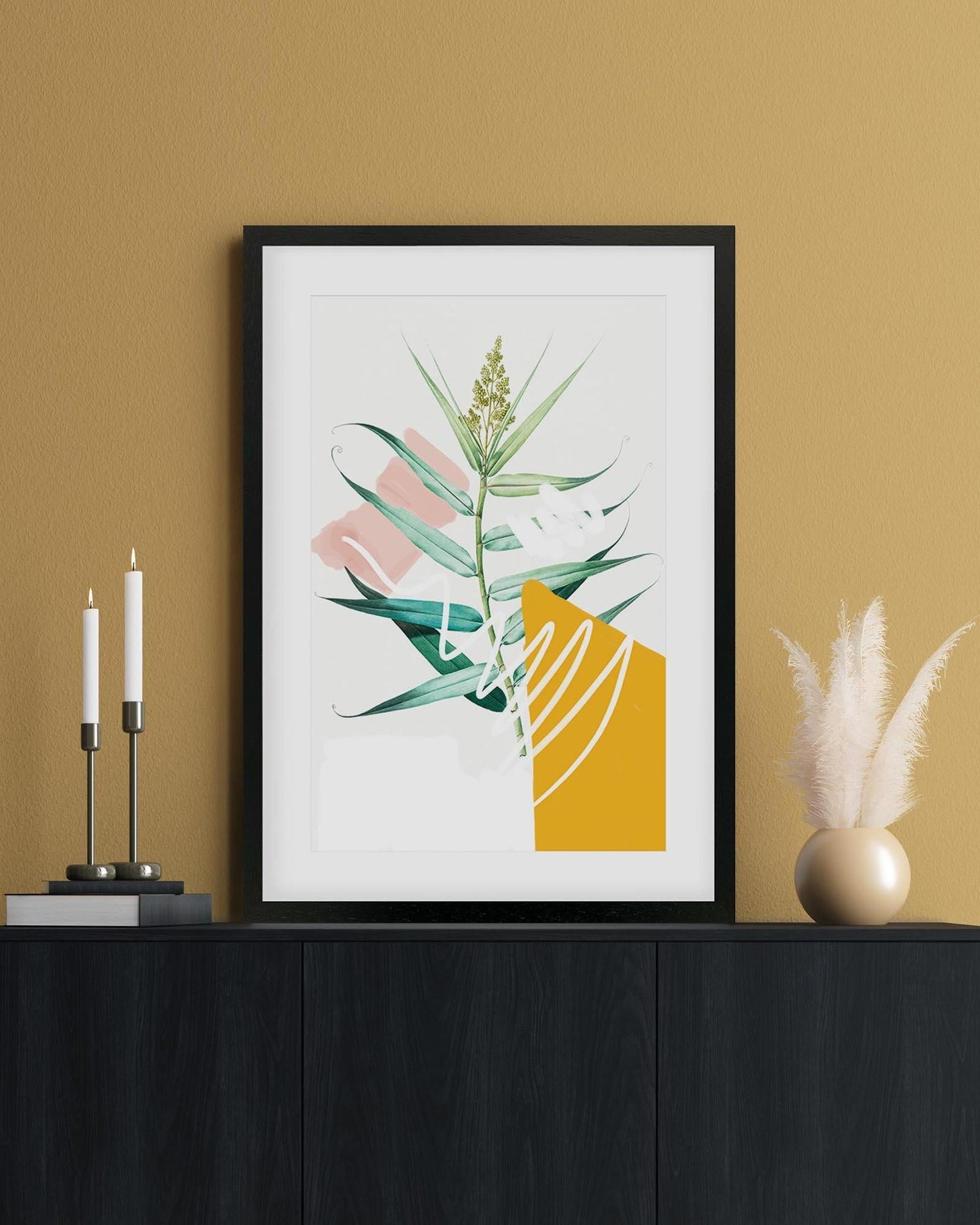 Framed abstract art print with green leaves and a yellow shape on a beige wall.