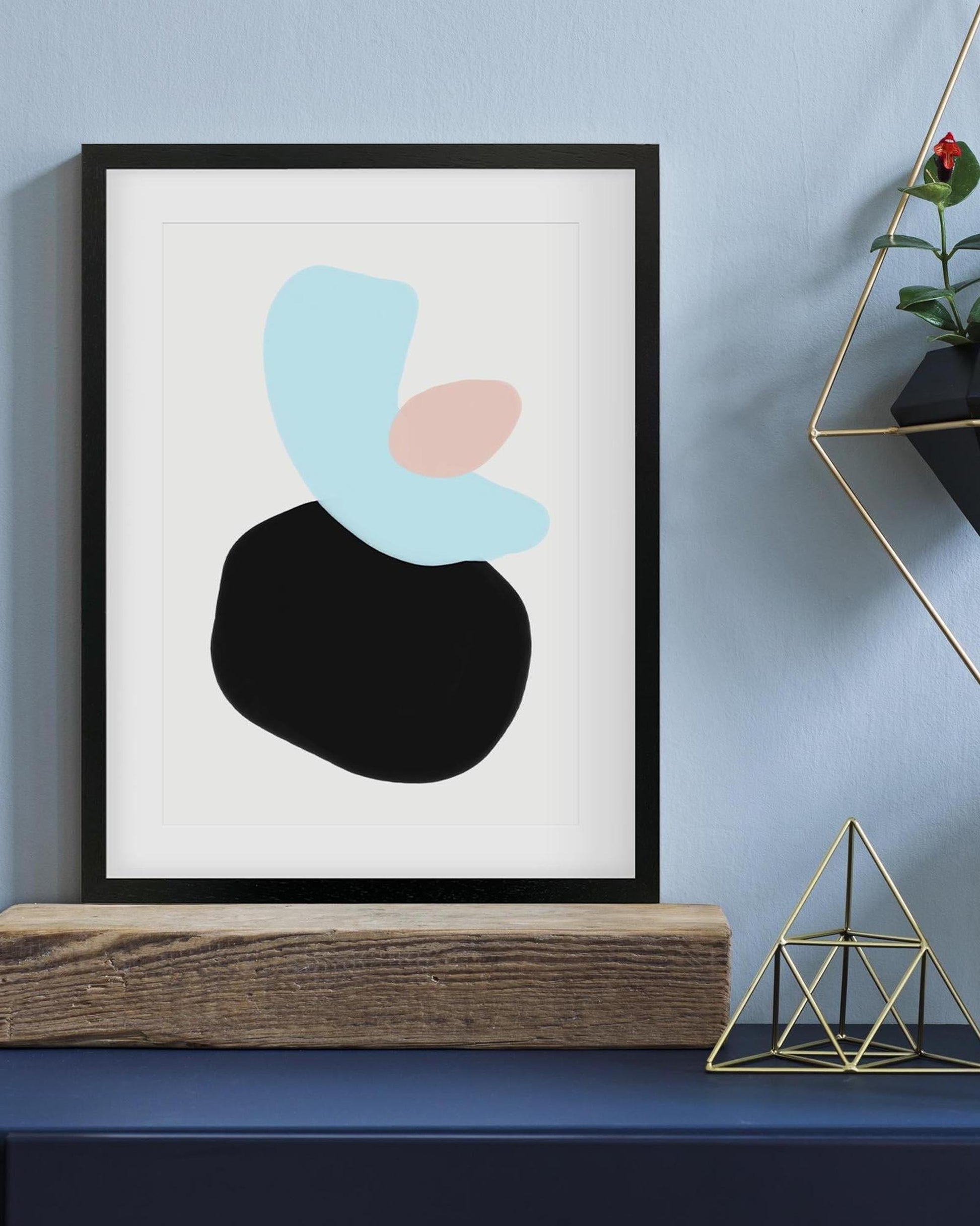 Abstract art print with geometric shapes in black, blue, and pink on a white background, framed in black against a light blue wall.