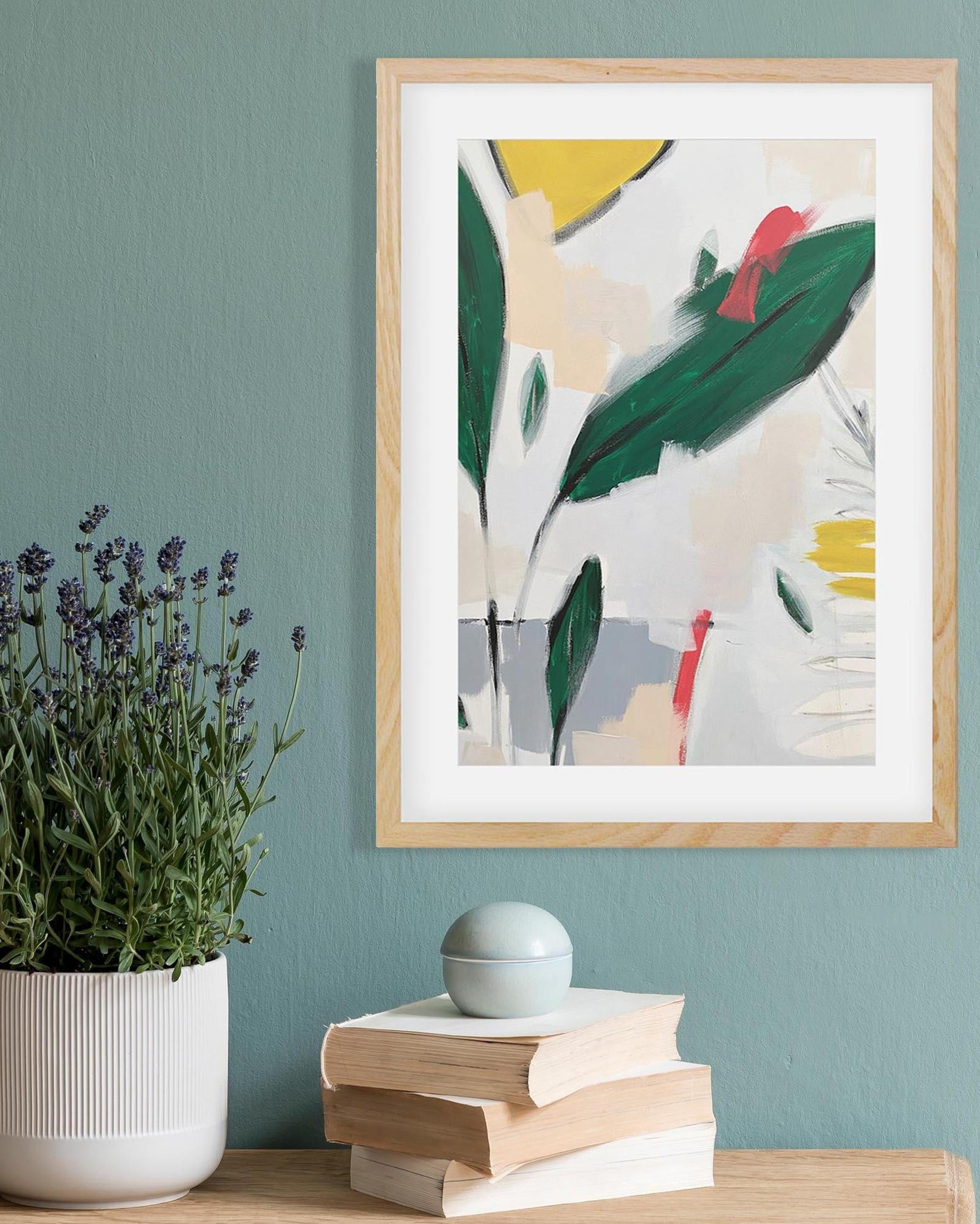Framed abstract art piece on a wall with books and a plant below.
