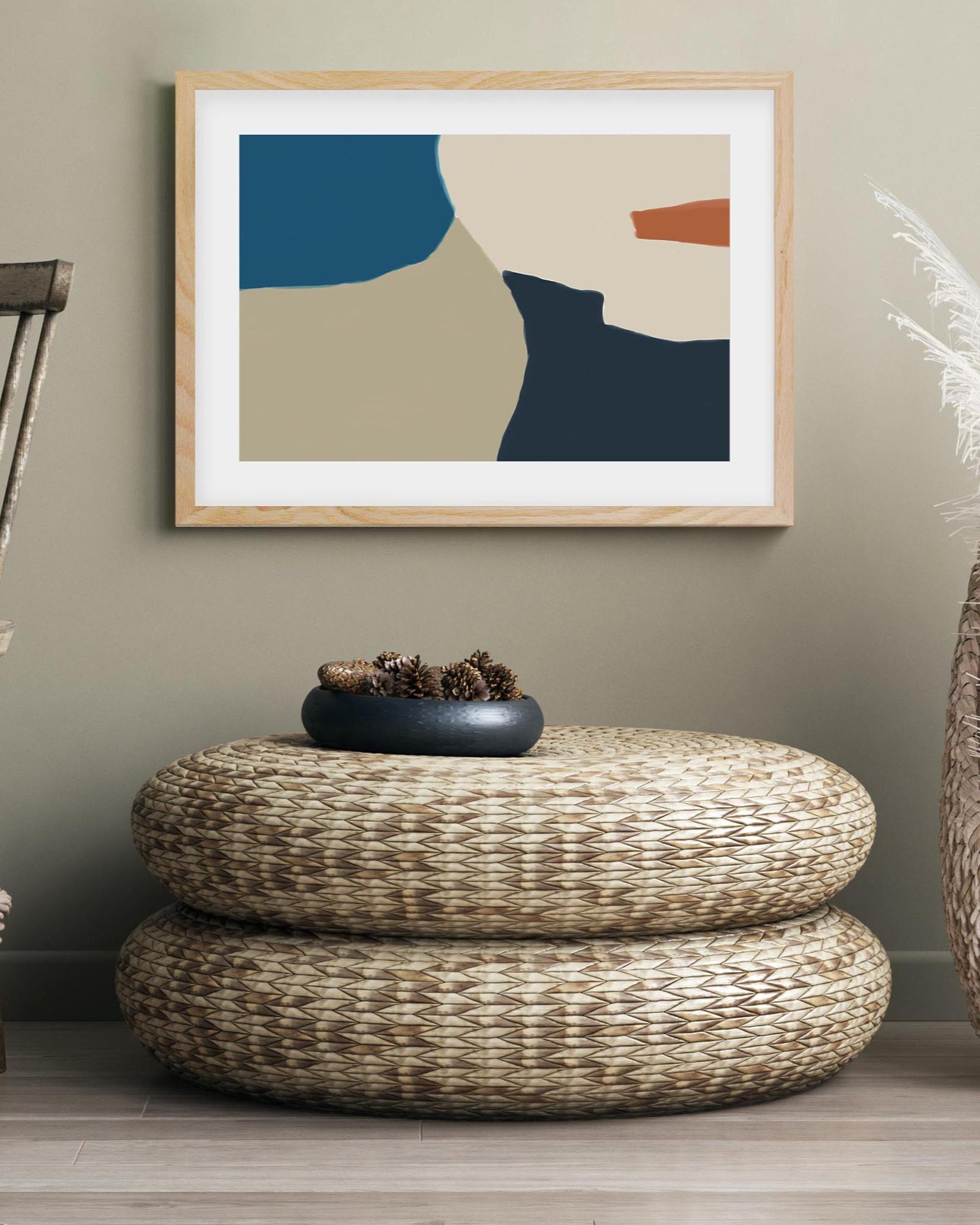 Abstract art print in a wooden frame on a wall above a woven ottoman.