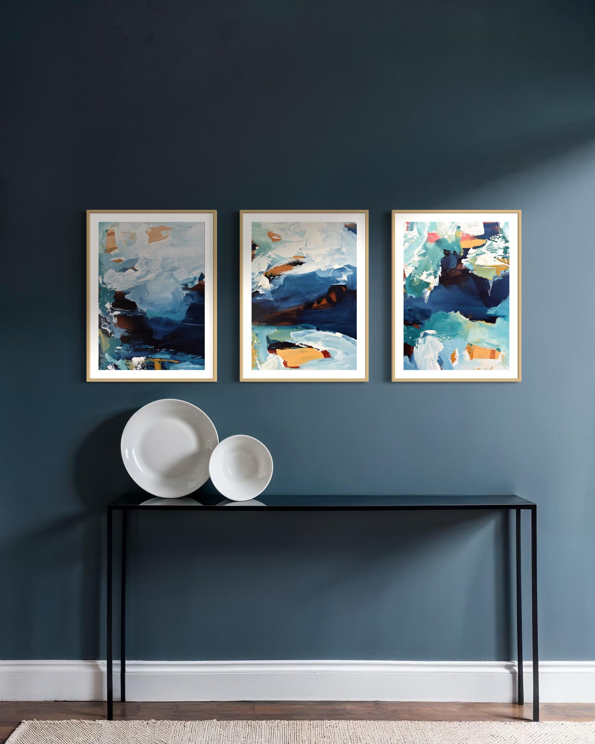 Three abstract art pieces on a dark blue wall above a black console table with white plates.